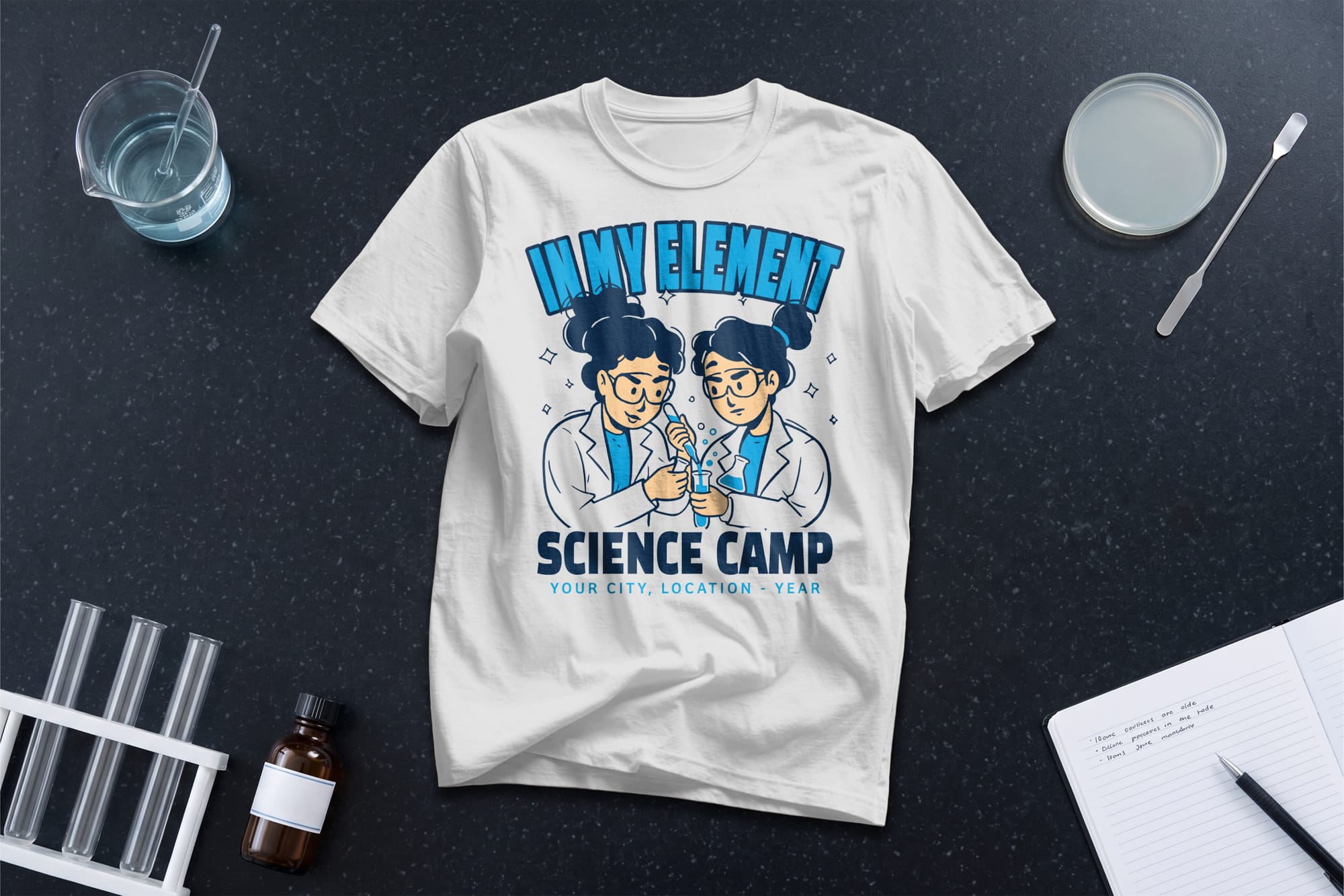 A white T-shirt with cartoon scientists and text "In My Element, Science Camp." Surrounding are lab tools and a notepad on a dark background.
