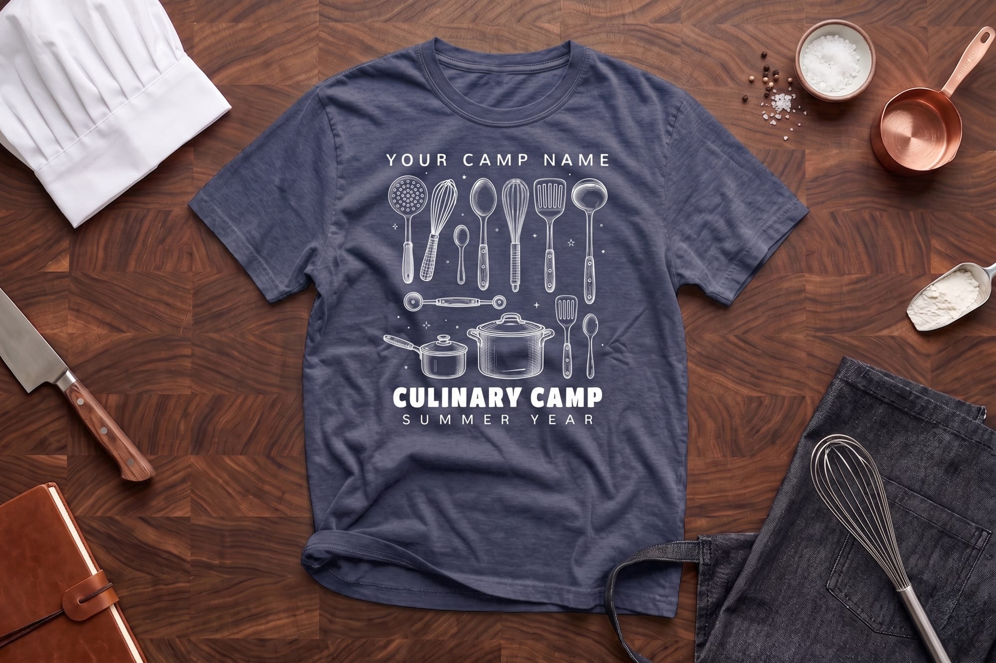 A blue T-shirt with culinary tool illustrations and "Culinary Camp" text lies on a wooden surface. Nearby are a chef hat, knife, apron, and copper measuring cup.