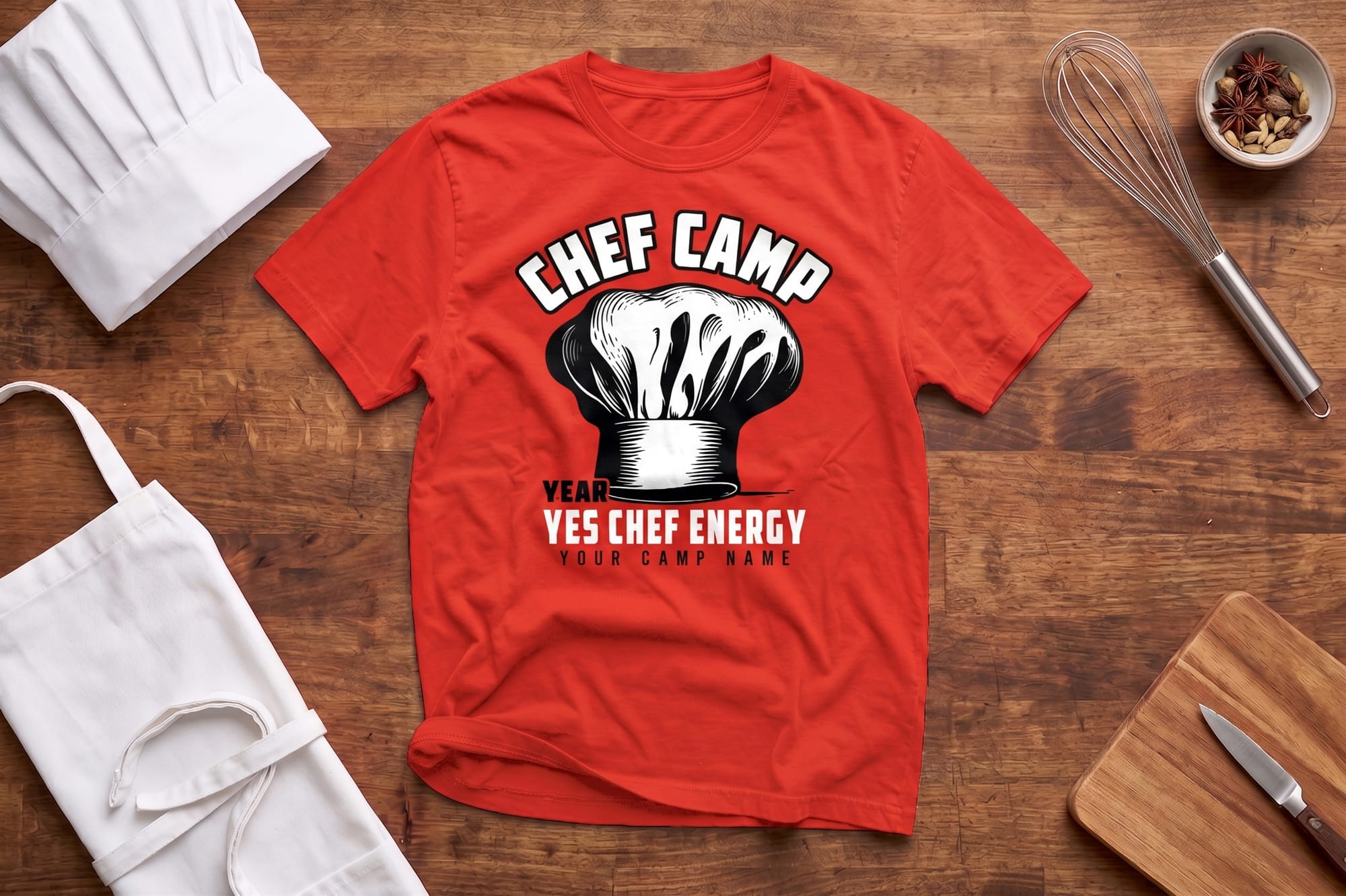 Red T-shirt with "Chef Camp" text and a chef hat graphic, placed on a wooden surface with a chef hat, apron, whisk, cutting board, and spices nearby.