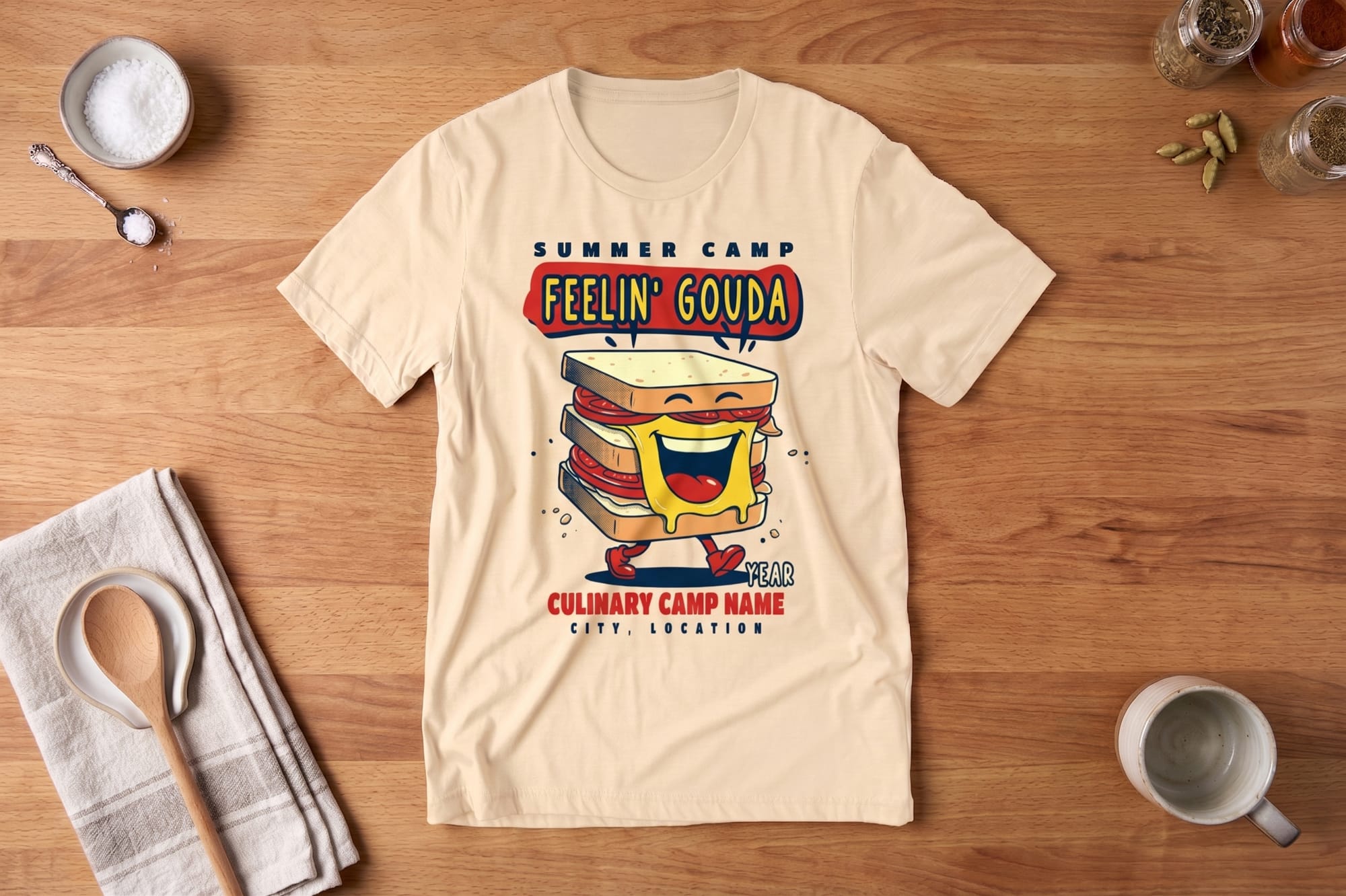 T-shirt on wood table with a cartoon sandwich character, smiling and melting cheese. Text: "Summer Camp Feelin' Gouda" and customizable camp details. Lighthearted tone.