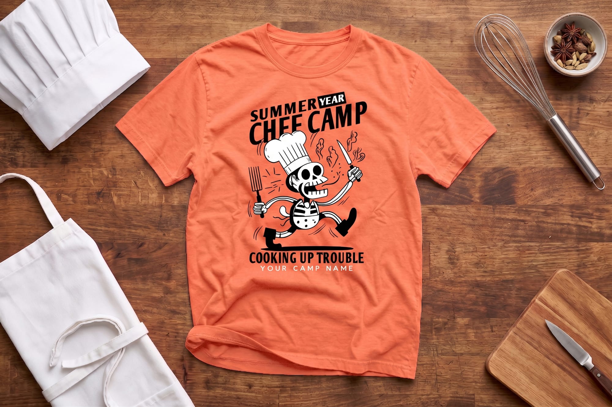 Orange T-shirt on a wooden surface with a cartoon skeleton chef design and text "Summer Year Chef Camp, Cooking Up Trouble." Nearby are a chef hat, apron, whisk, and spices.
