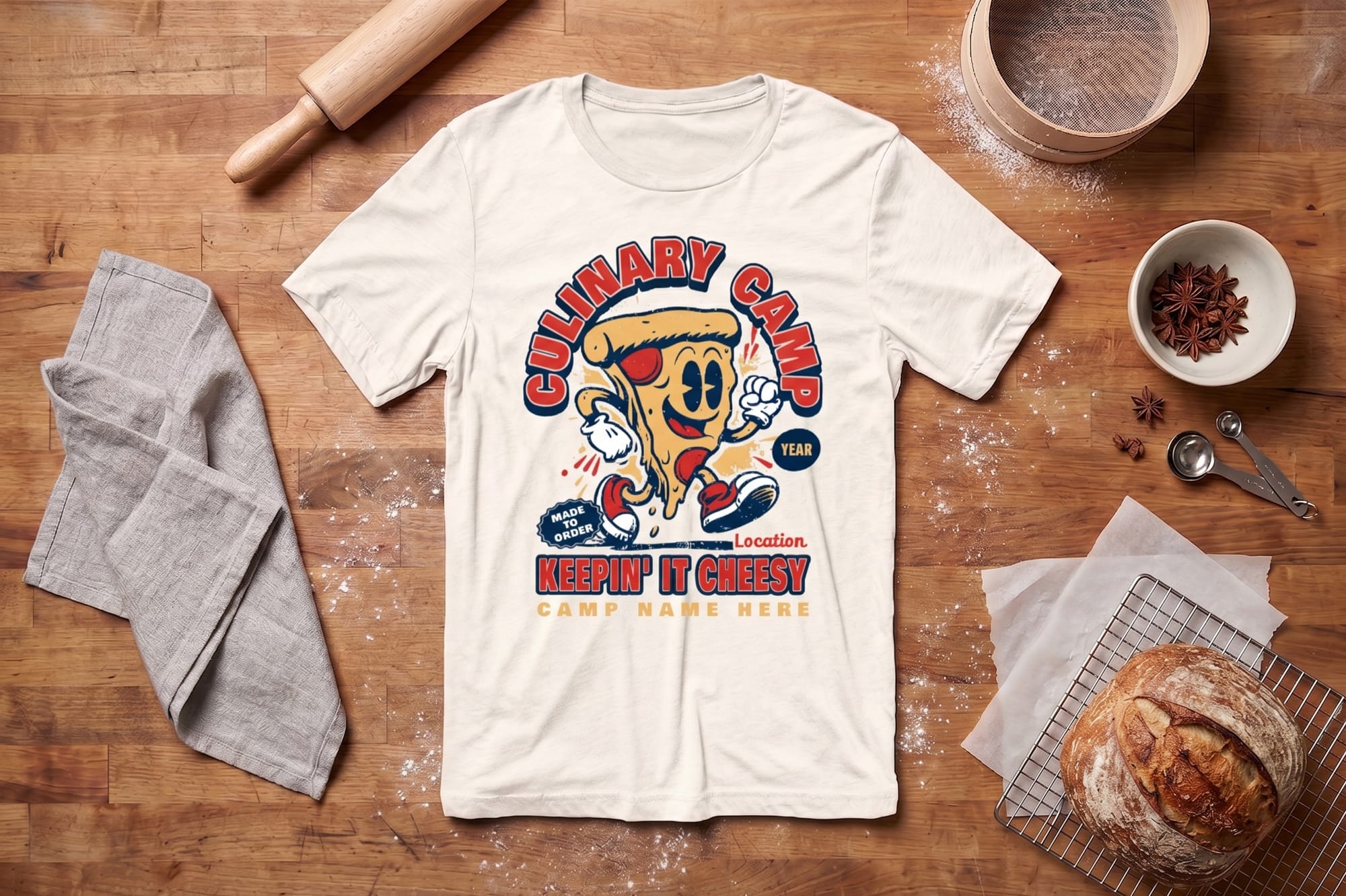 T-shirt with a colorful, retro pizza slice cartoon saying "Culinary Camp: Keepin' It Cheesy." It lies on a wooden surface surrounded by baking items: a rolling pin, gray towel, bread, and spices, evoking a fun, culinary vibe.