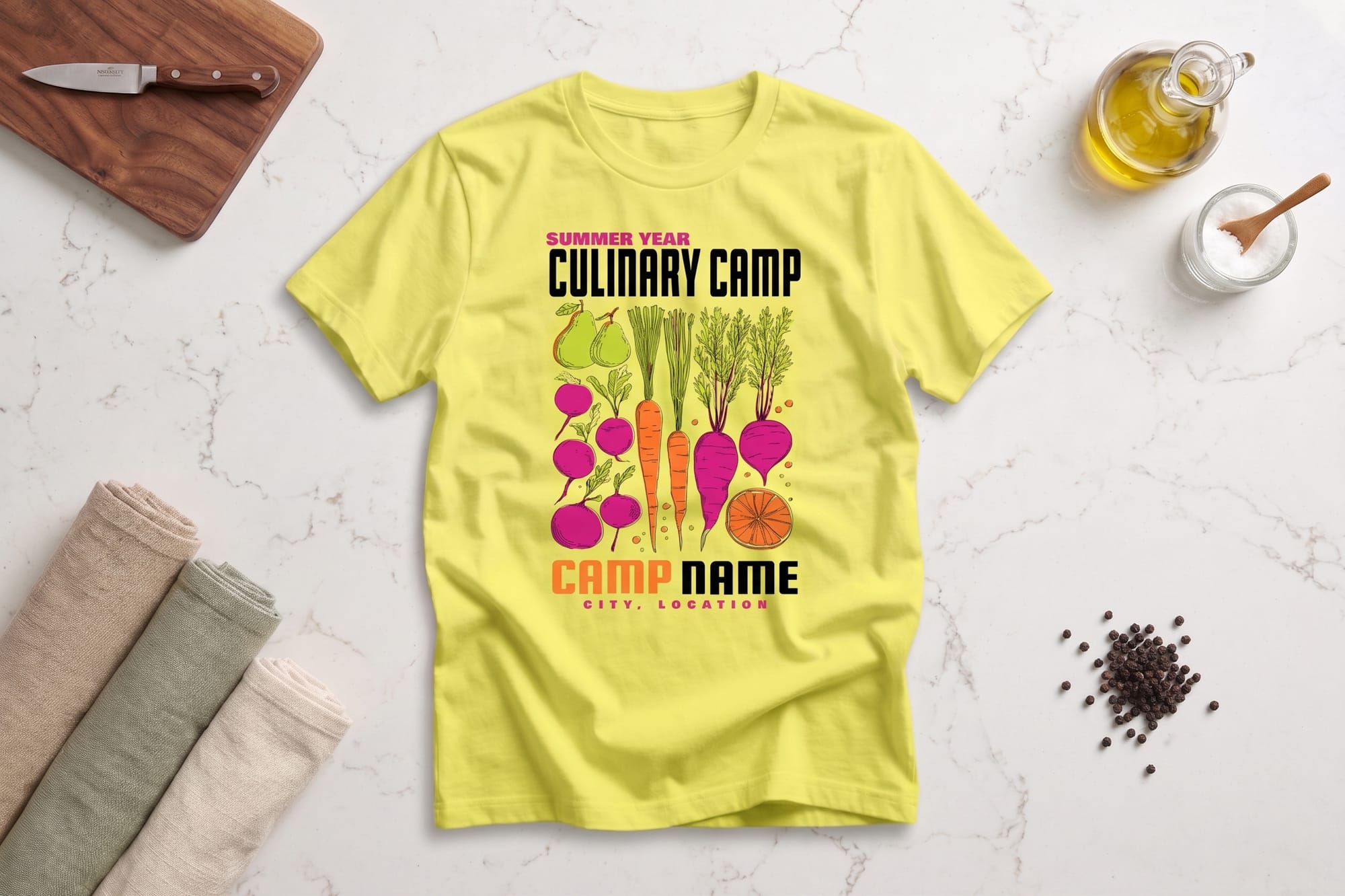 Yellow t-shirt on marble table with the text "Summer Year Culinary Camp" and colorful vegetables. Nearby are towels, a cutting board, and kitchen items.