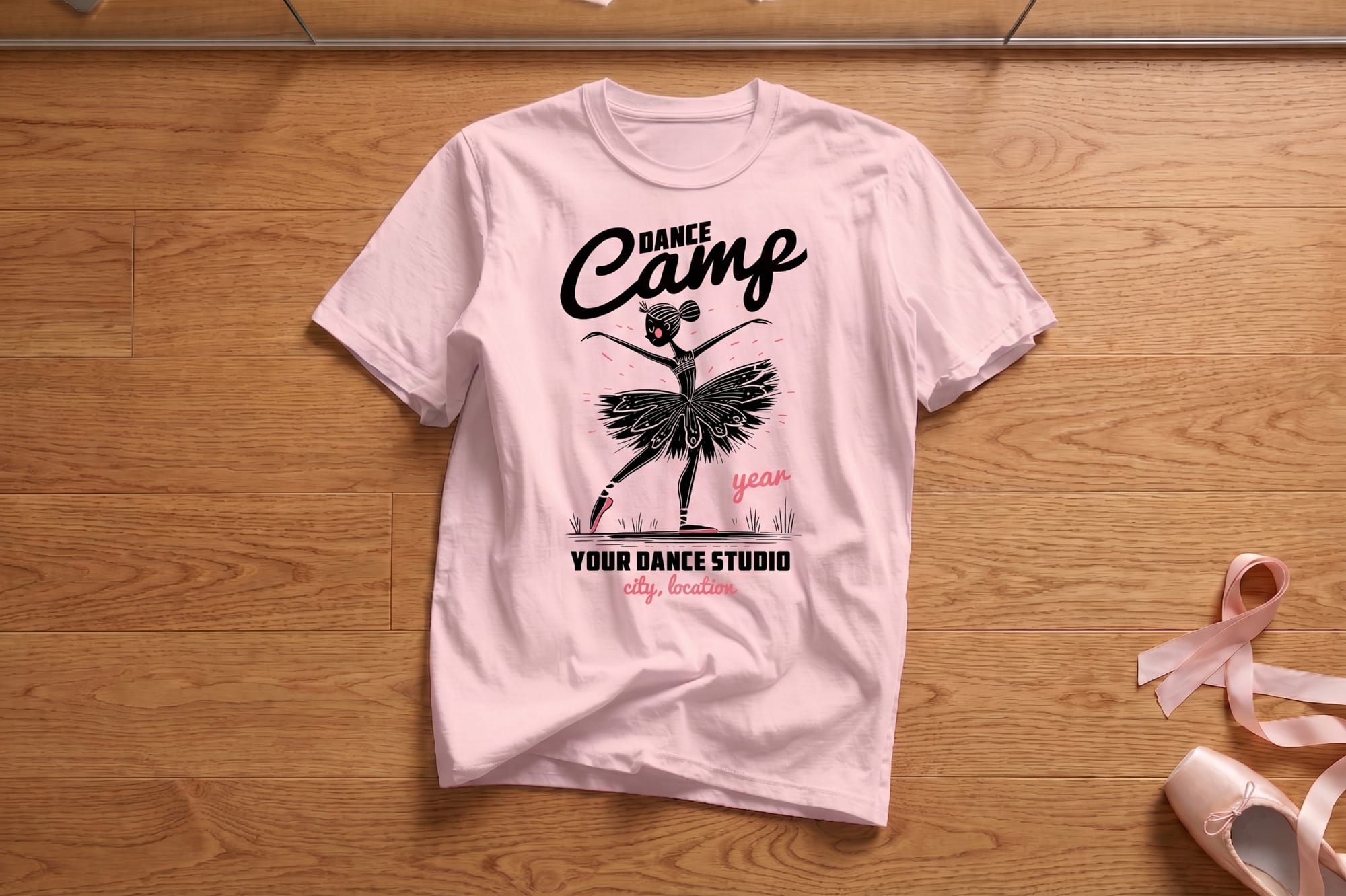 Pink t-shirt on wooden floor, featuring a black-and-white ballerina illustration with text "Dance Camp" and customizable studio details. Ballet shoes nearby.