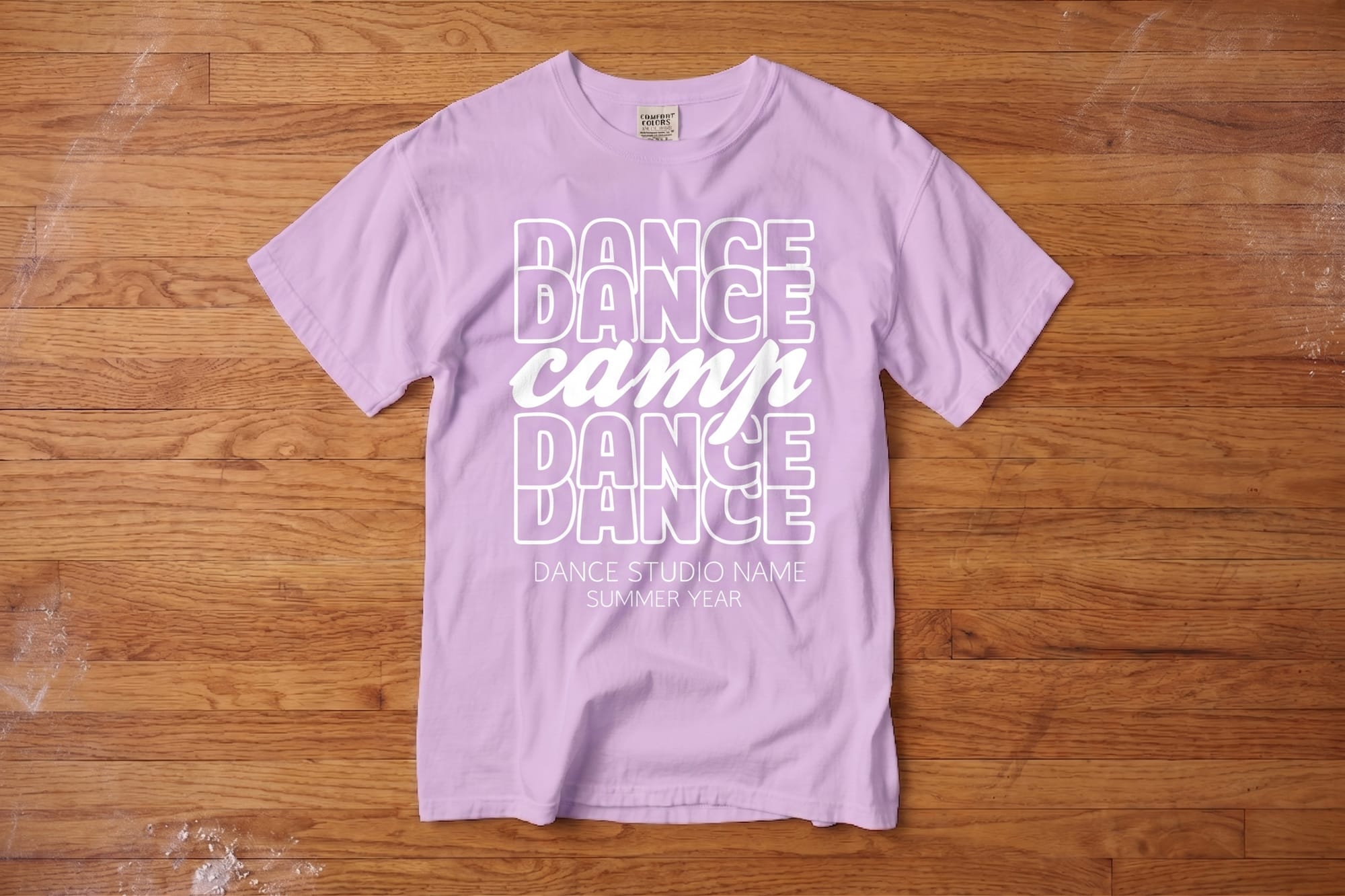 Lavender T-shirt on wood floor, featuring bold white text reading “DANCE CAMP DANCE” with “CAMP” in script. Below: "Dance Studio Name, Summer Year."