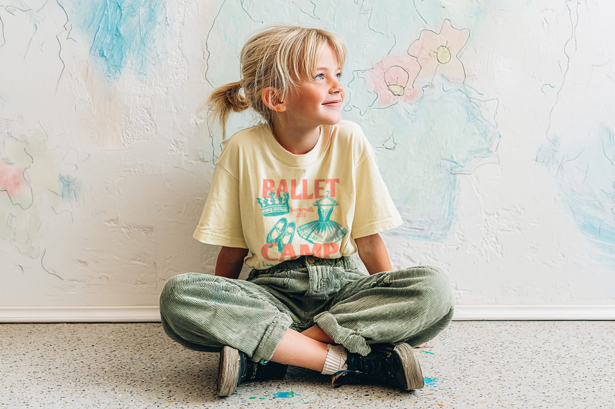 A smiling child in a "Ballet Camp" shirt sits cross-legged against a pastel-colored wall with hand-drawn flowers, exuding a joyful, playful vibe.