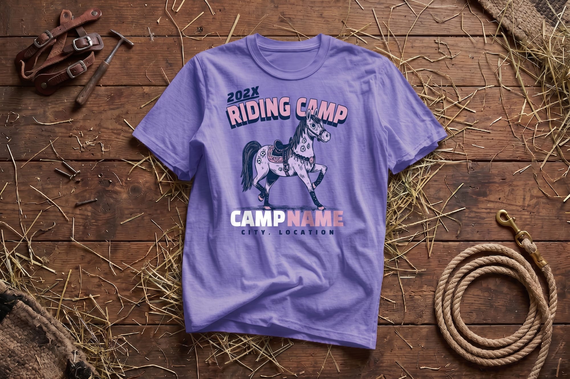 Lilac t-shirt with "202X Riding Camp" graphic featuring a horse, placed on a wooden surface. Surrounding items include hay, a coiled rope, and leather gear, creating a rustic, equestrian theme.