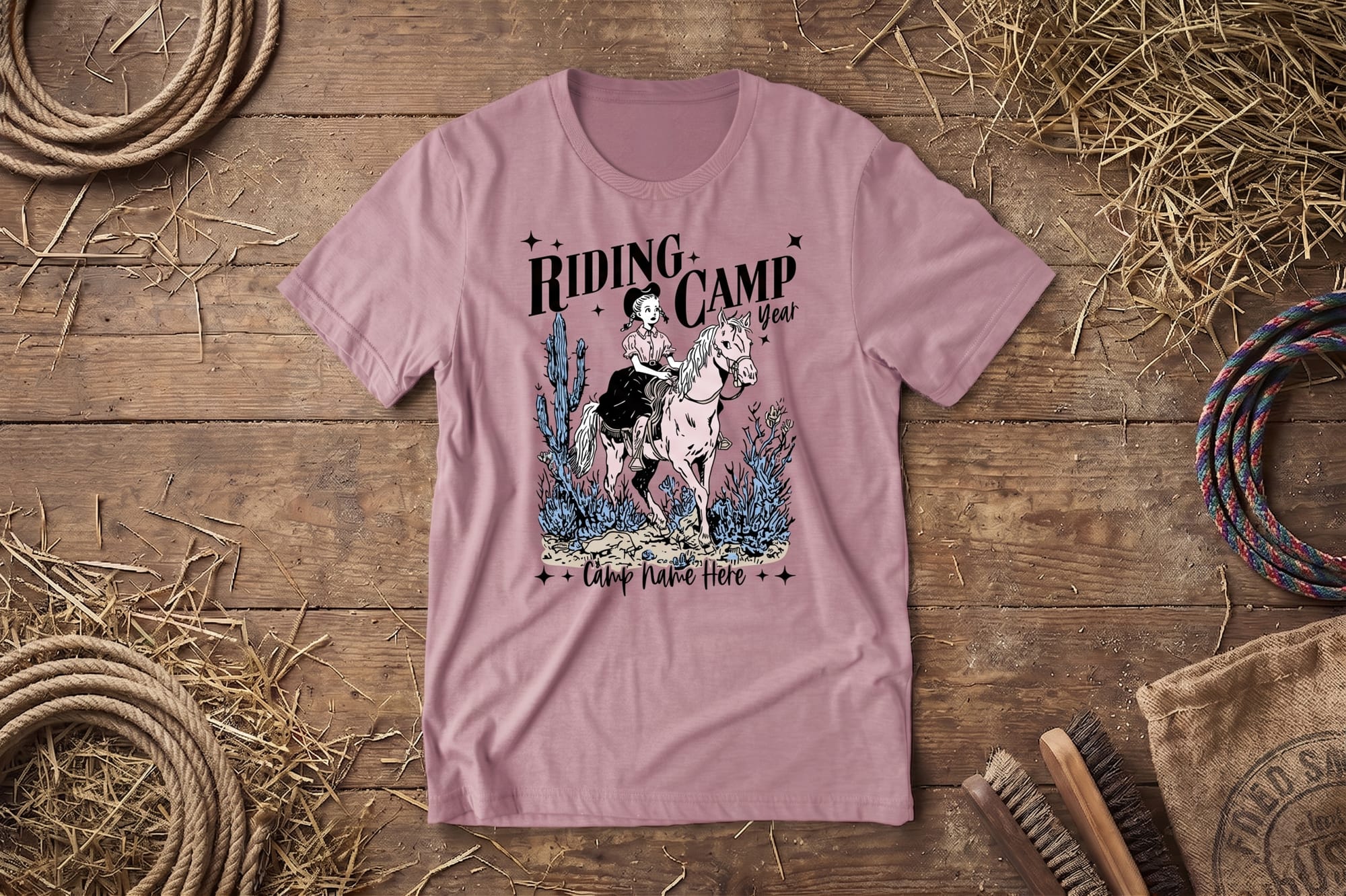 T-shirt on a wooden surface, featuring an illustration of a person riding a horse, surrounded by plants. Text reads "Riding Camp" and "Camp Name Here." Rustic elements include ropes and hay, conveying a country theme.