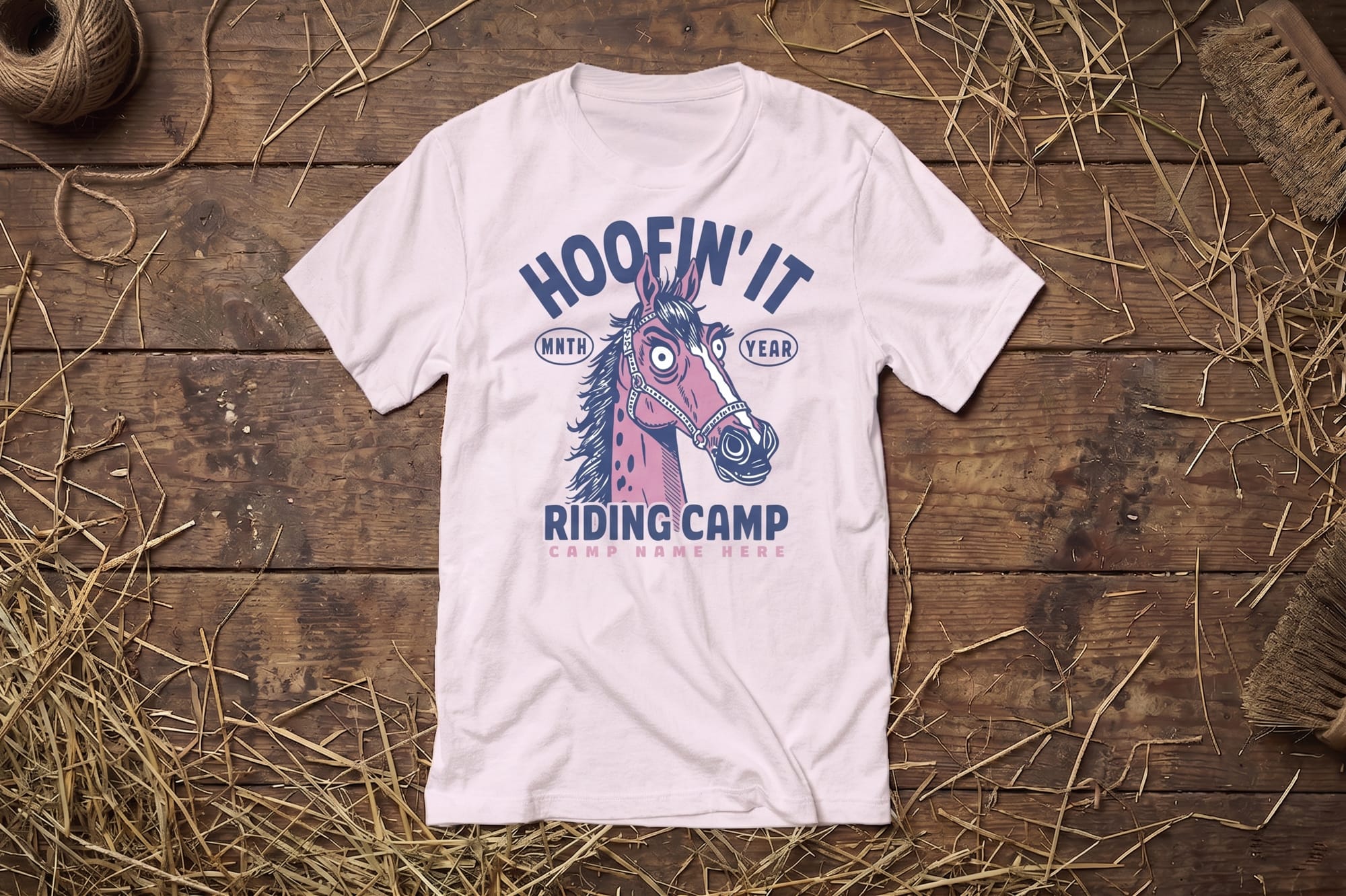 Light lilac T-shirt on wooden floor with hay, featuring a cartoon horse and text "Hoofin' It Riding Camp." Rustic and playful equestrian theme.