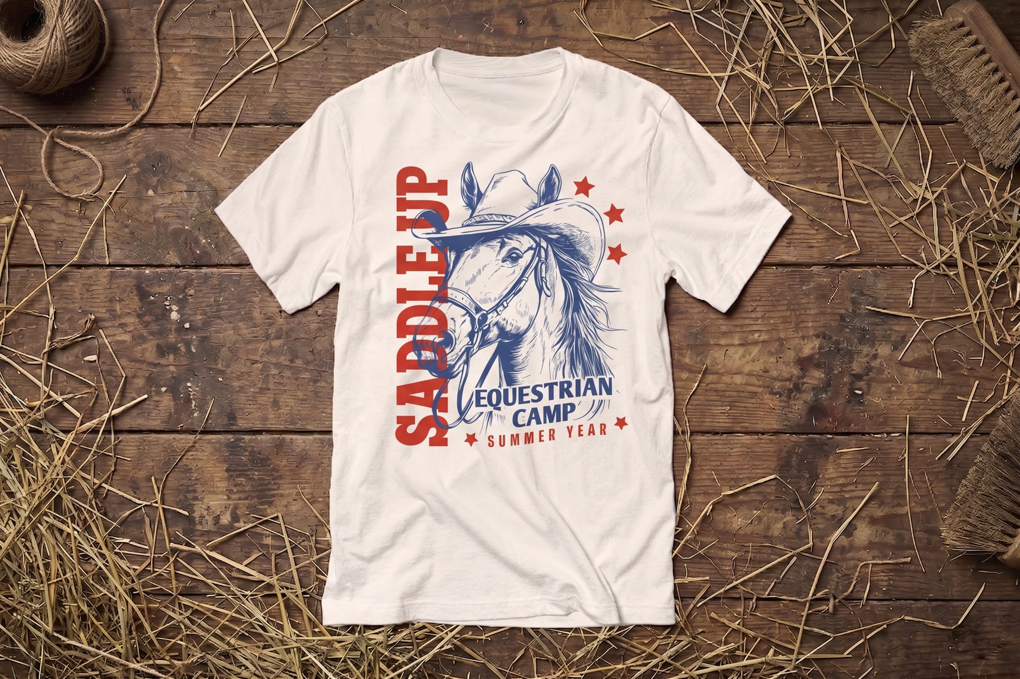 T-shirt on rustic wooden floor with hay and rope. Design features a horse in a cowboy hat, red stars, and text "Saddle Up Equestrian Camp."