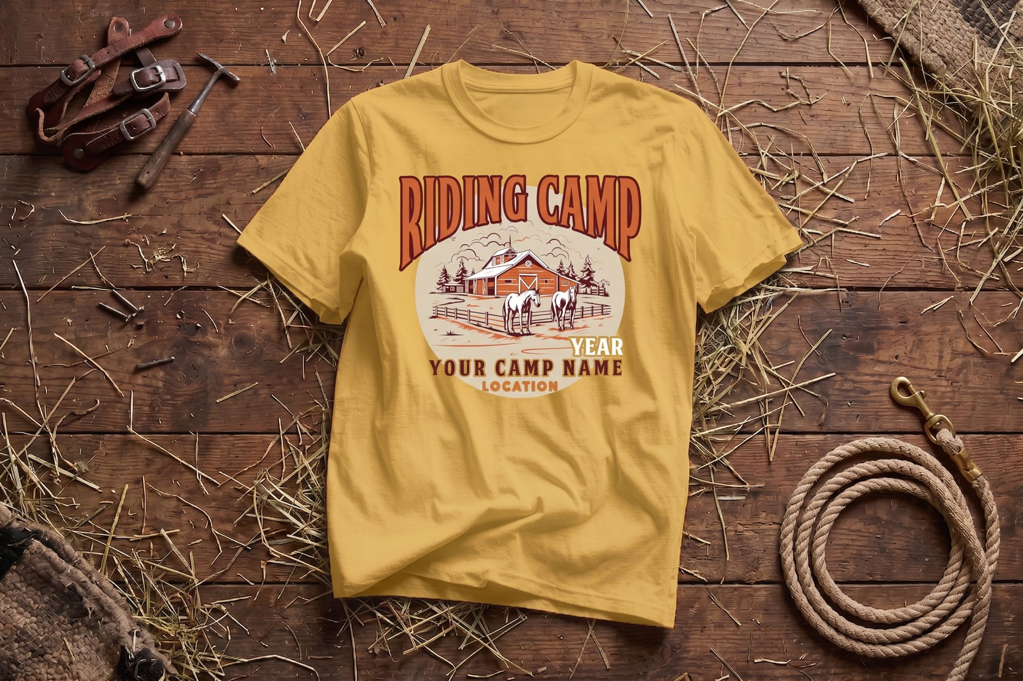 Yellow T-shirt on wooden floor, printed with "Riding Camp" and a farm scene. Surrounding items include a coiled rope, leather straps, and hay, creating a rustic feel.