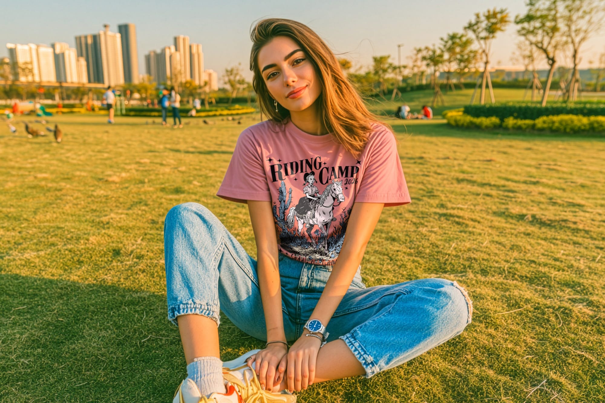 Young woman sitting cross-legged on grass in a park, wearing a pink "Riding Camp" T-shirt and jeans. Cityscape in background, warm sunset lighting.