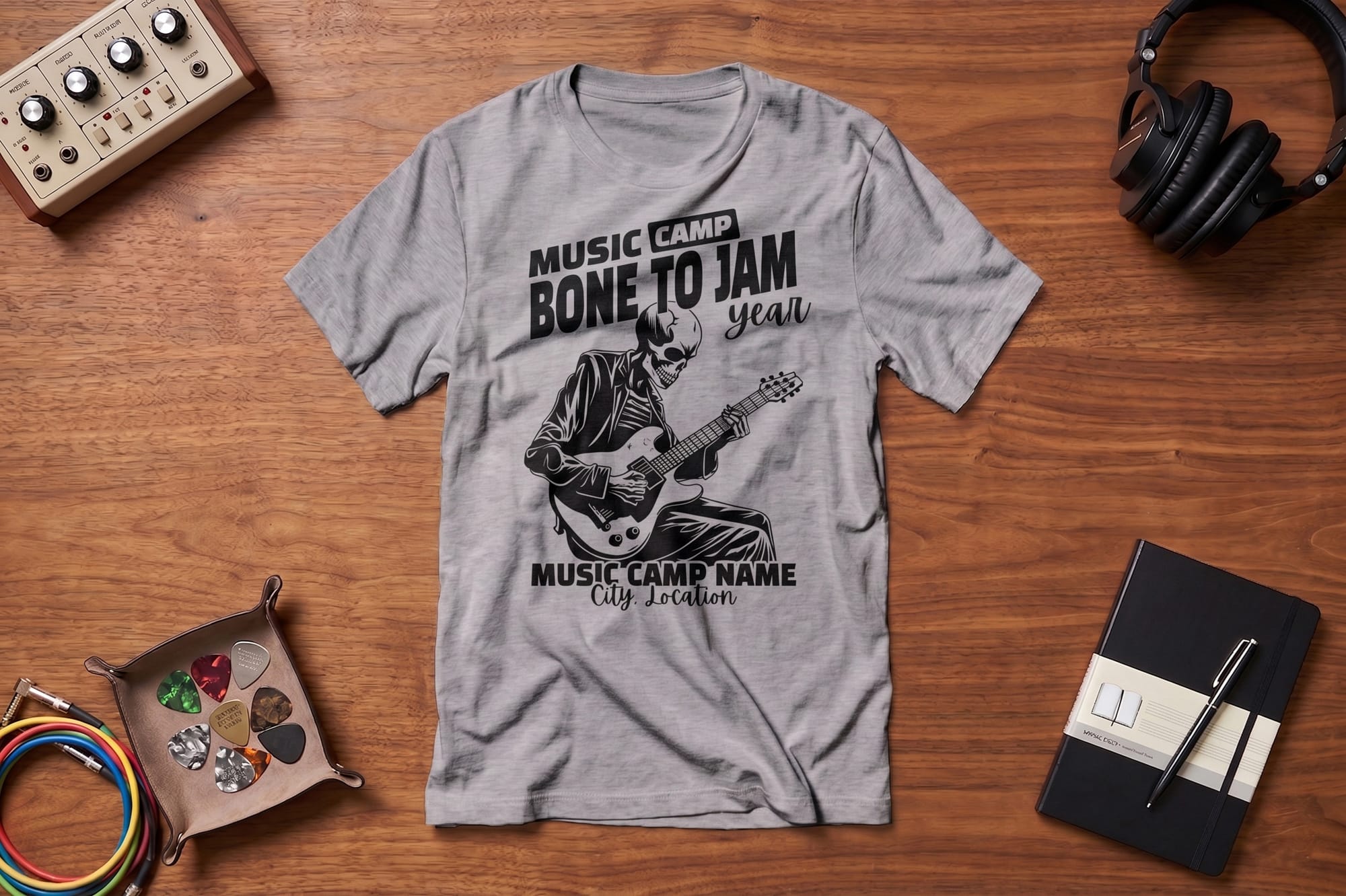 Gray t-shirt with "Music Camp Bone to Jam" design featuring a skeleton playing guitar. Surrounding items: guitar picks, cables, notebook, headphones.