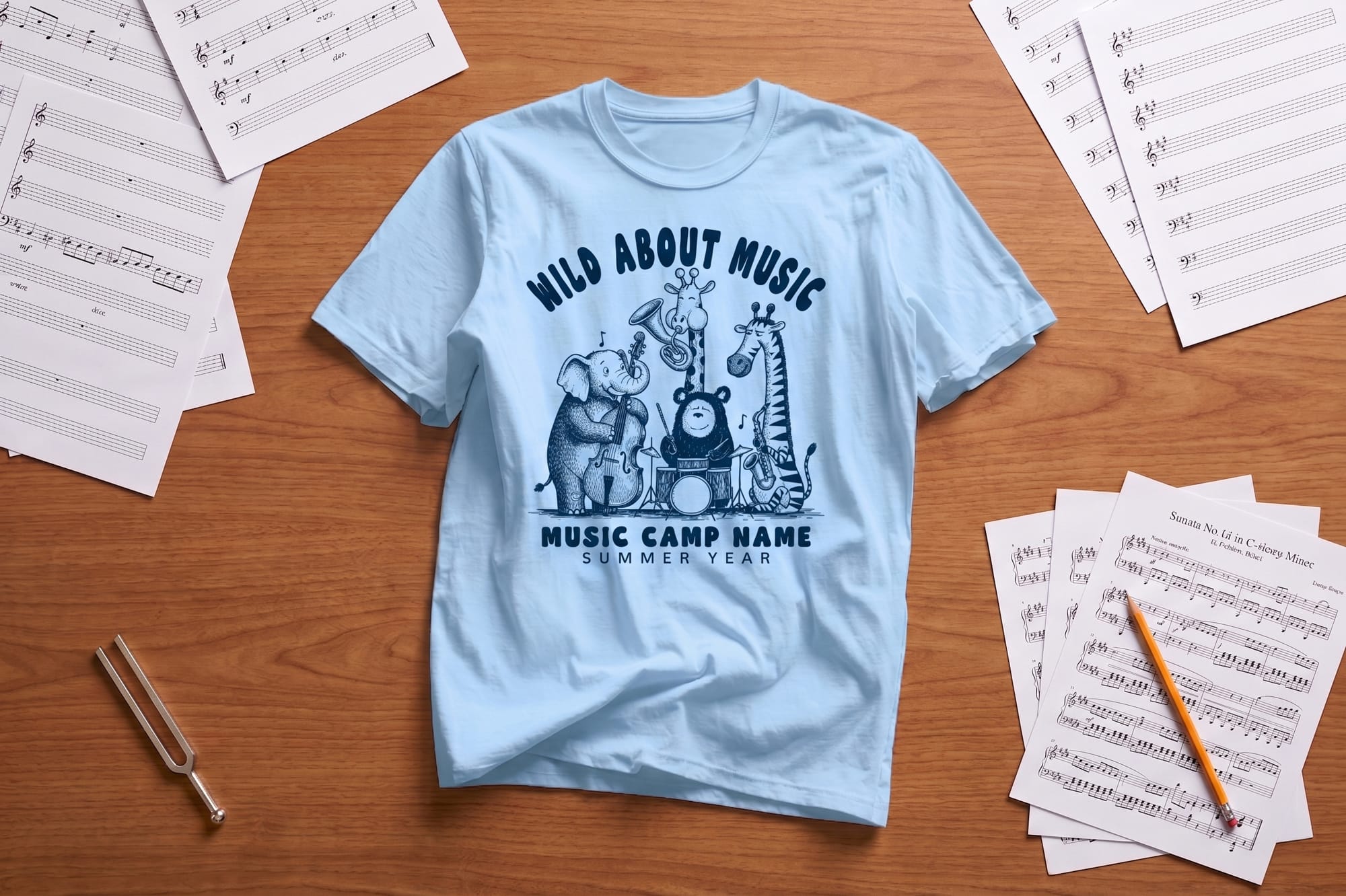 Light blue T-shirt with "Wild About Music" design featuring cartoon animals playing instruments. Surrounded by sheet music, pencil, and tuning fork on a wooden table.