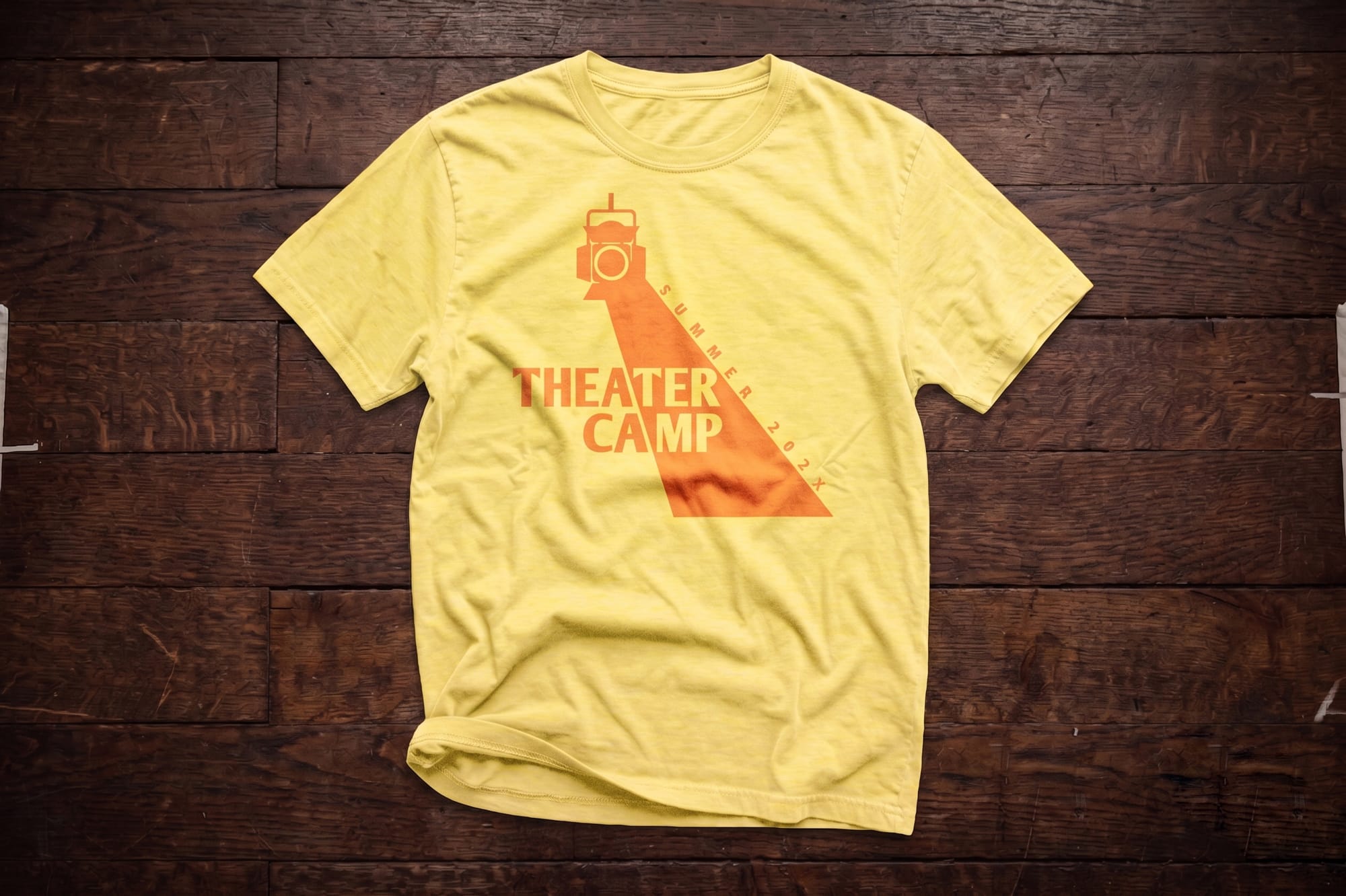 Yellow crewneck t-shirt with an orange stage spotlight graphic and 'Theater Camp Summer 202X' text, flatlay on wood