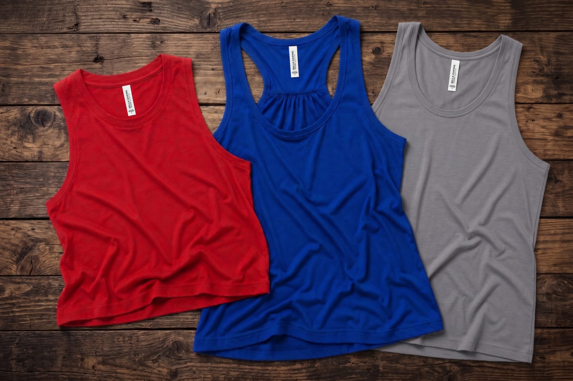 Three blank Bella+Canvas tank tops in red, royal blue racerback, and gray, laid flat on a wood surface