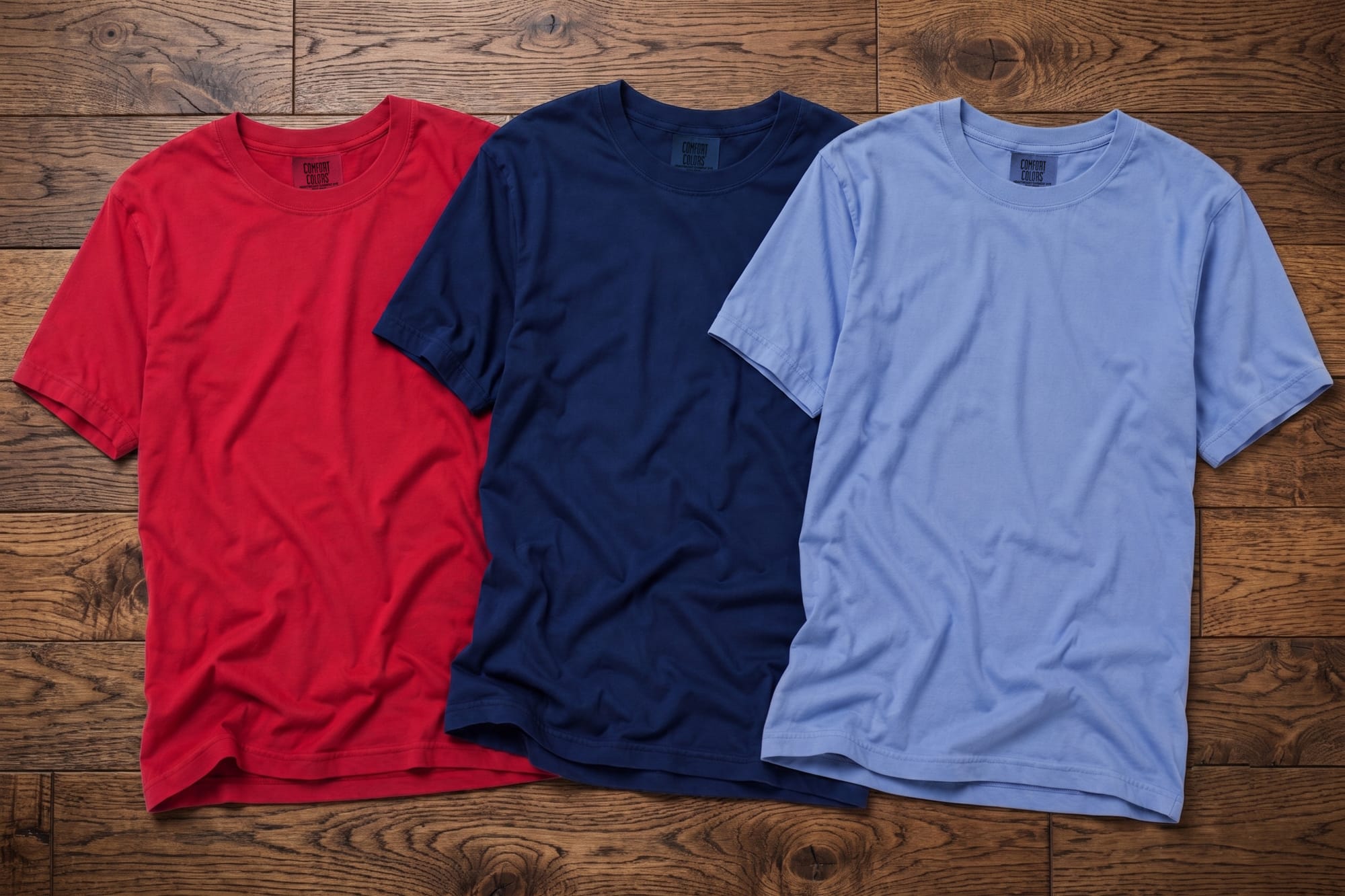 Three blank Comfort Colors crewneck t-shirts in red, navy, and light blue laid flat on a wood surface