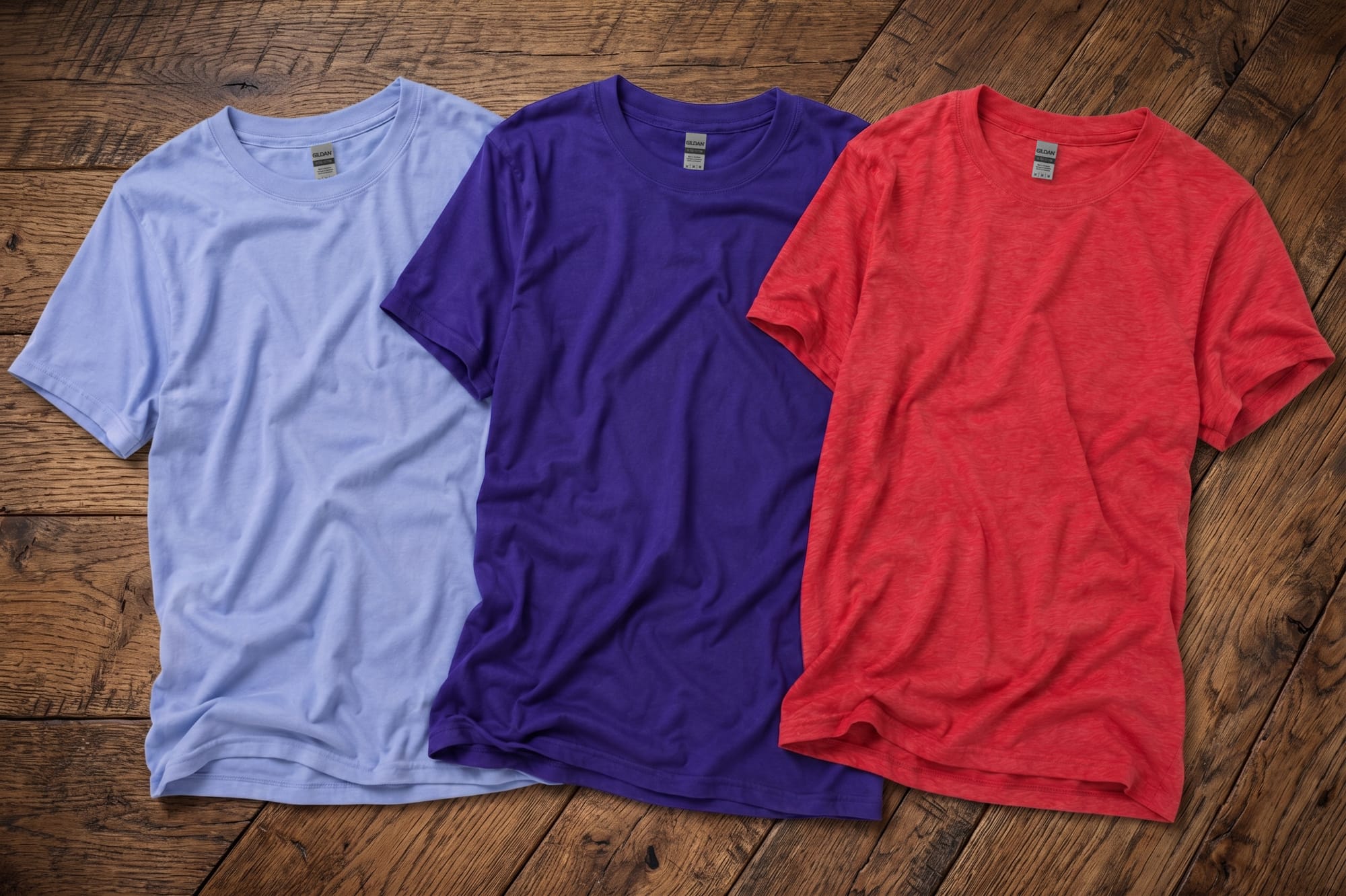 Three blank crewneck t-shirts in light blue, navy, and heather red laid flat on a wood surface