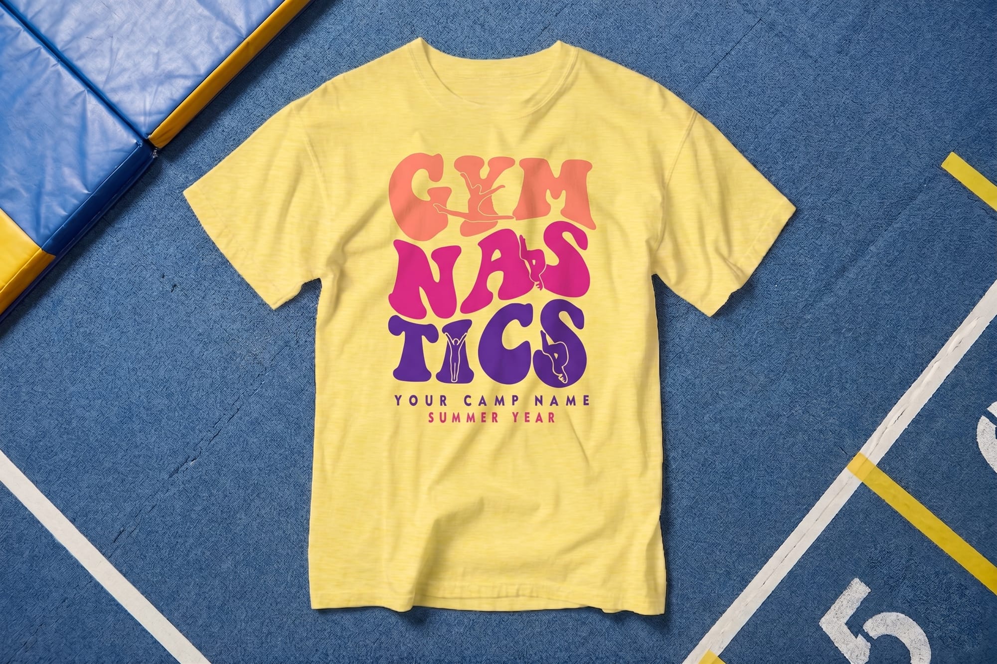 Yellow crewneck t-shirt with a retro-style custom-printed 'Gymnastics' design in coral, pink, and purple, laid flat on a gymnasium floor mat