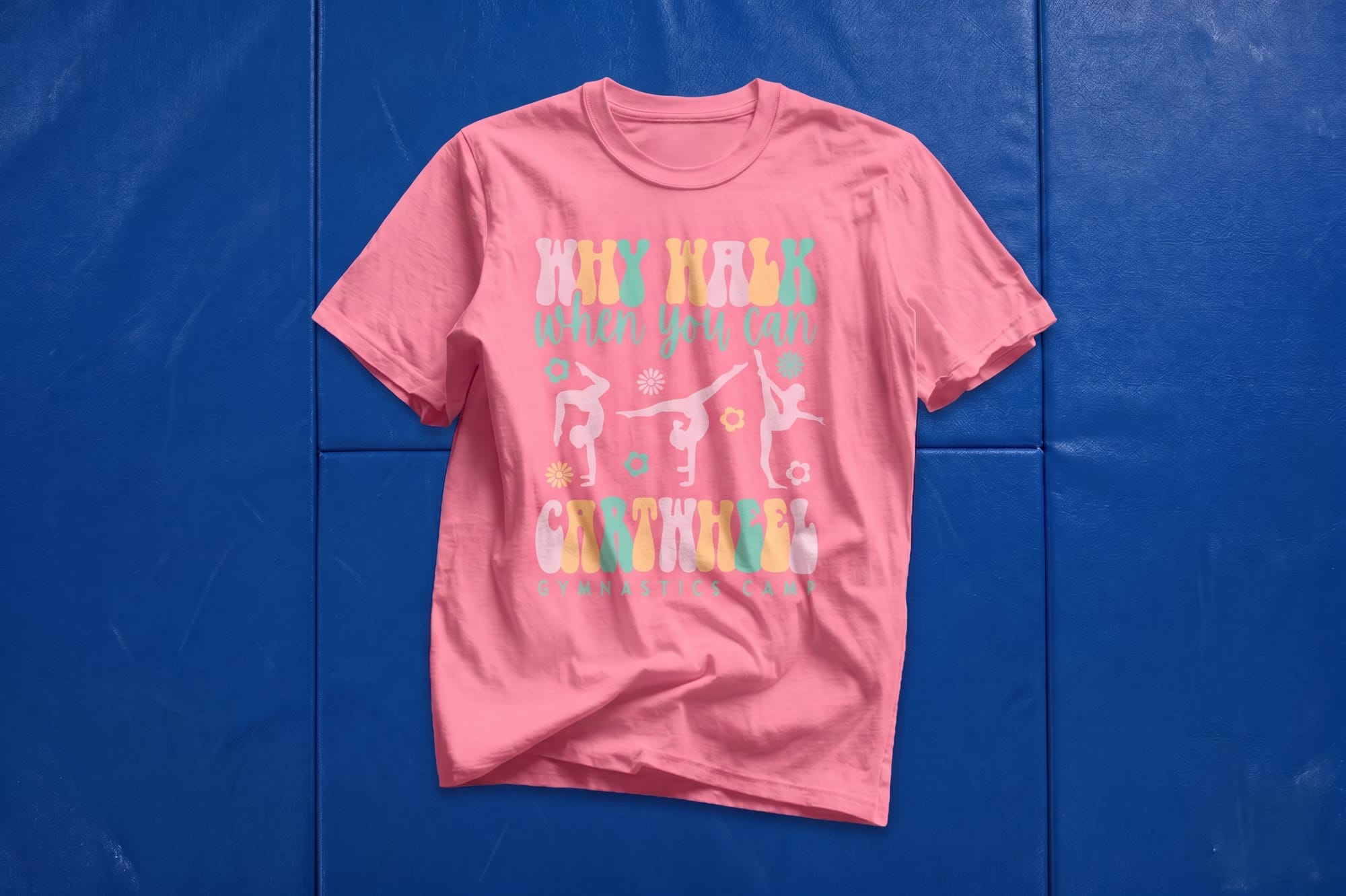 Pink crewneck t-shirt with retro custom print reading 'Why Walk When You Can Cartwheel' with gymnast silhouettes, laid flat on a blue gymnastics mat