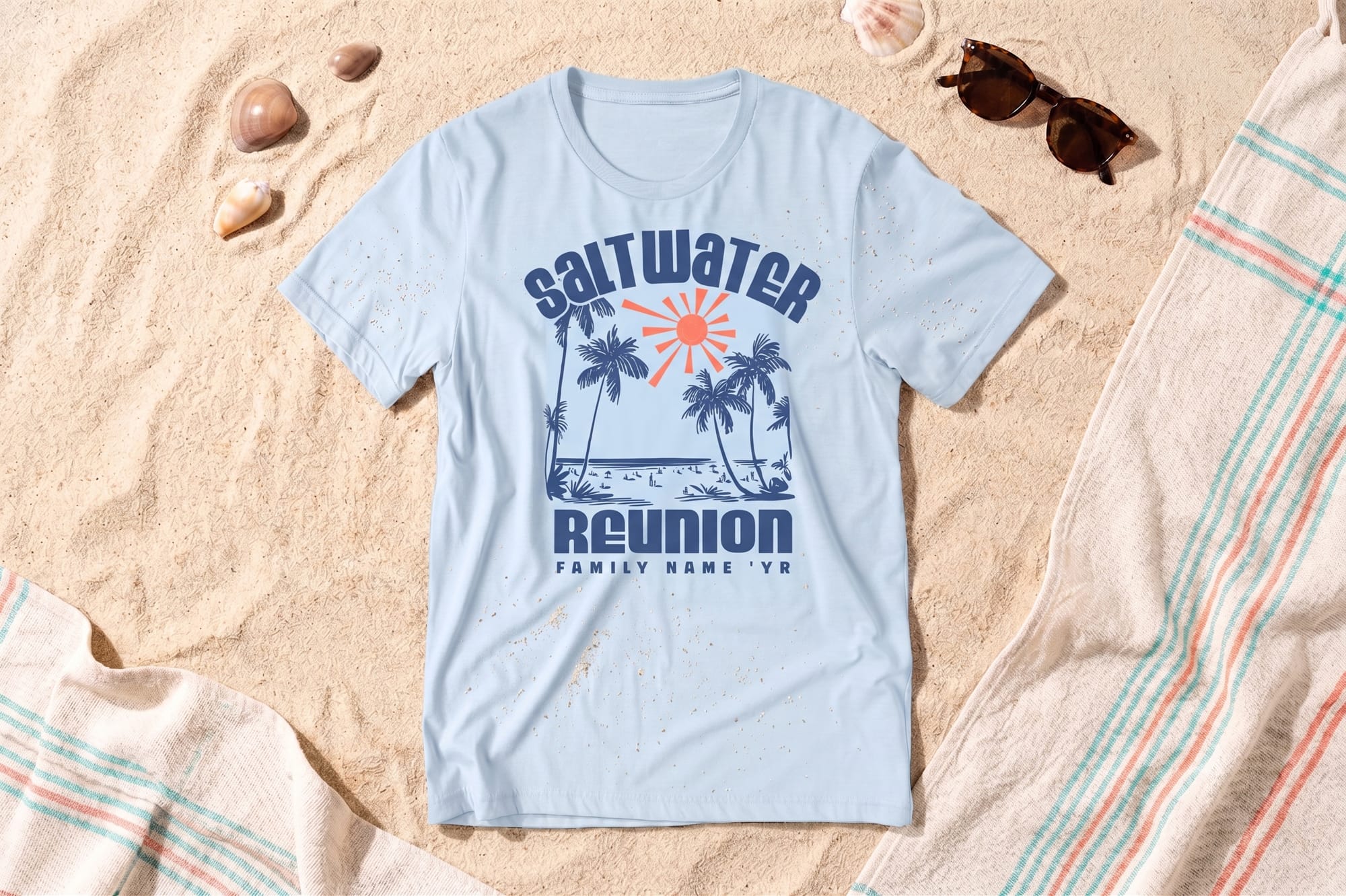 Light blue crewneck tee with a retro beach screen-printed design reading 'Saltwater Reunion' with palm trees and a sun, laid flat on sand