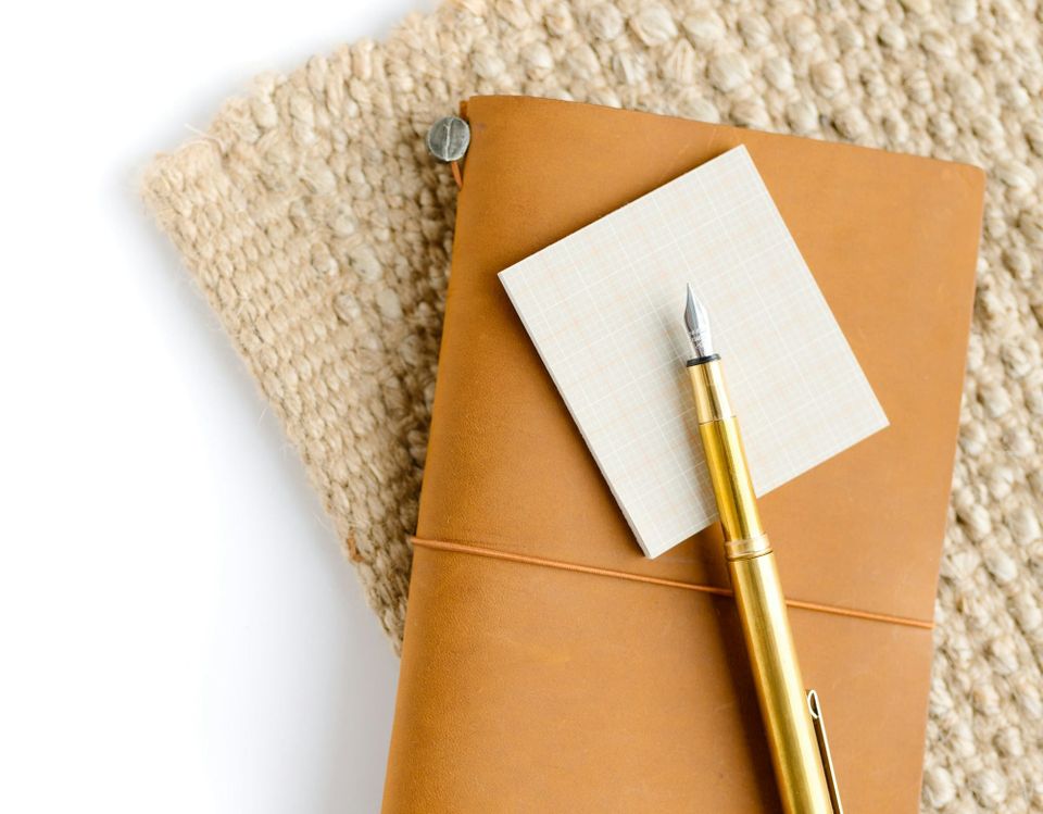 Photo of a light tan leather Traveler's Notebook cover laying on a jute fabric placemat, with a brass fountain pen on top.