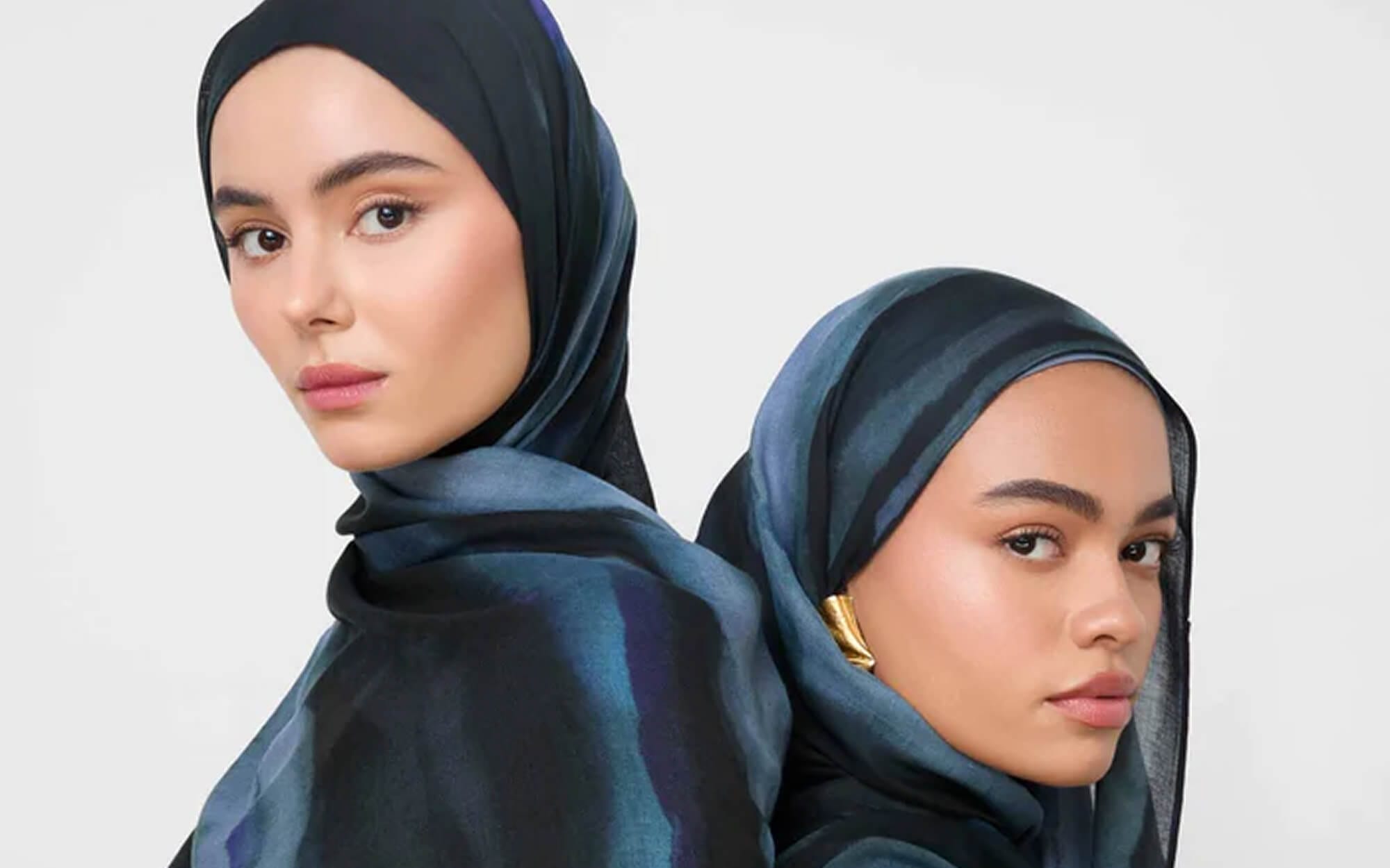 Two women wearing bold, dark teal and navy patterned hijabs styled neatly around their faces, standing back-to-back against a neutral background.