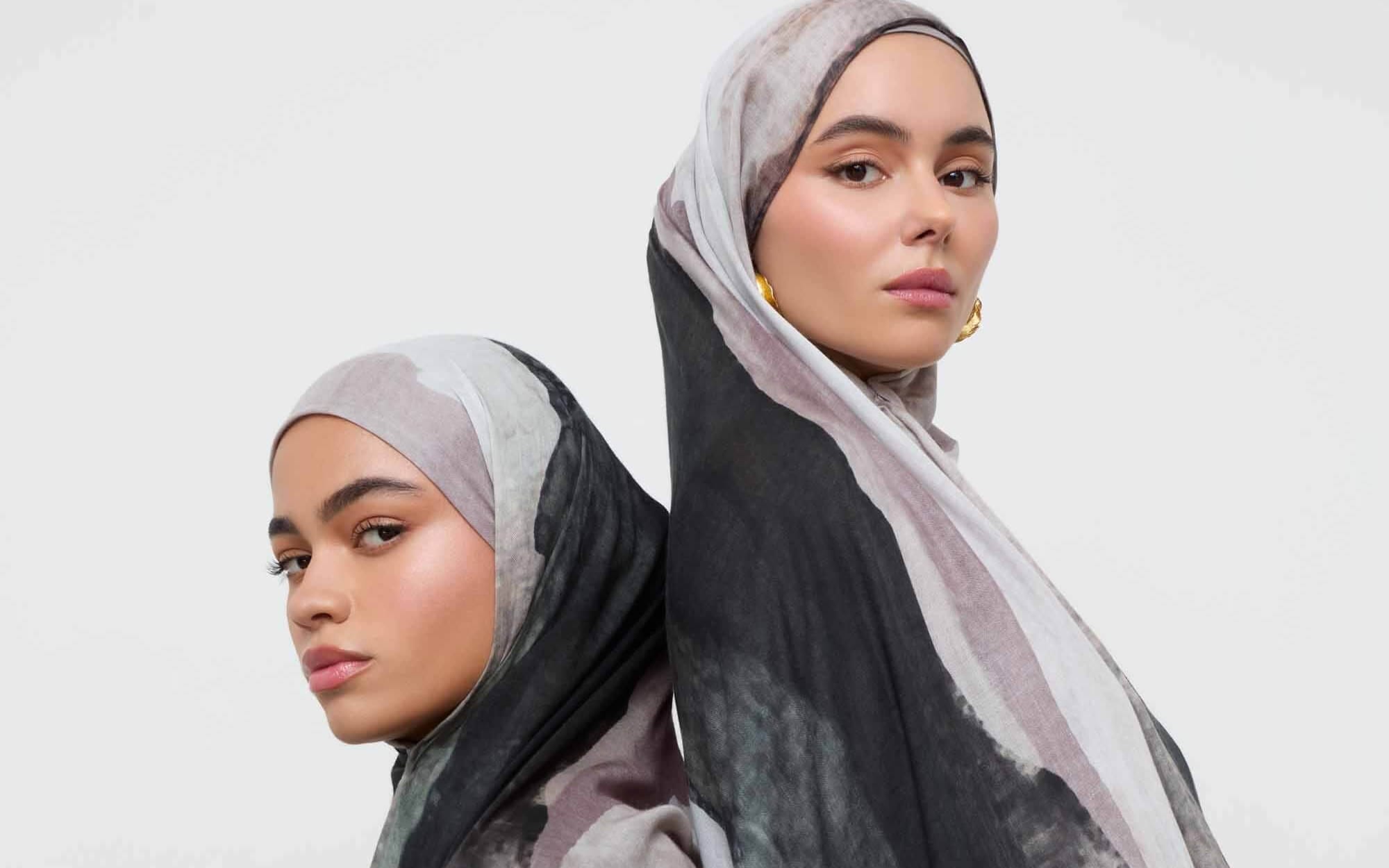 Two women wearing soft marble-print hijabs in muted grey, black, and lavender tones, standing back-to-back against a plain light background.