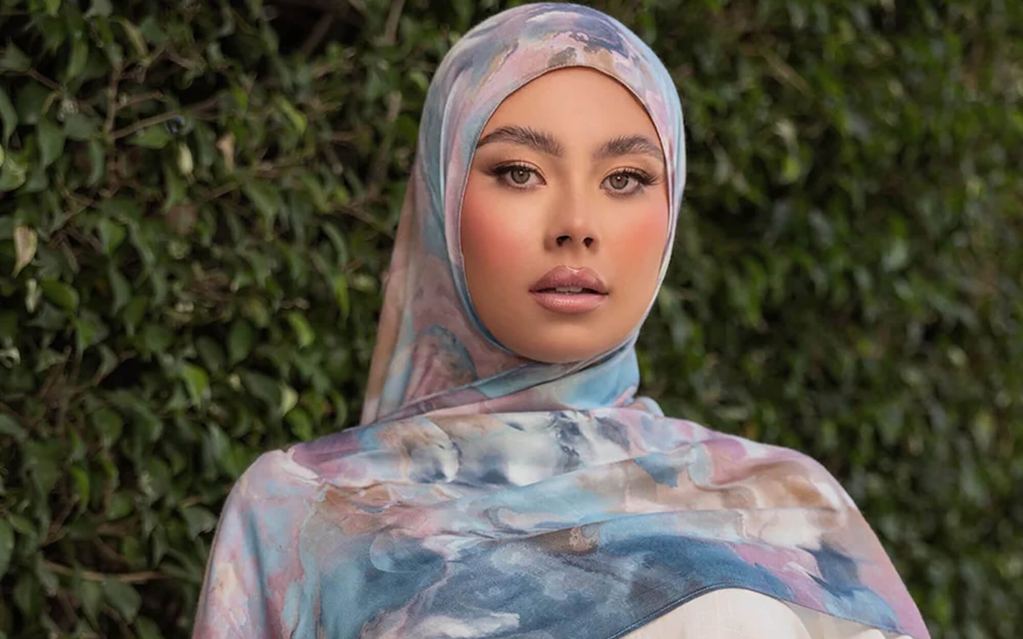 A woman wearing a soft marble-print hijab in pastel blue, pink, and lavender tones, standing in front of a green leafy background.