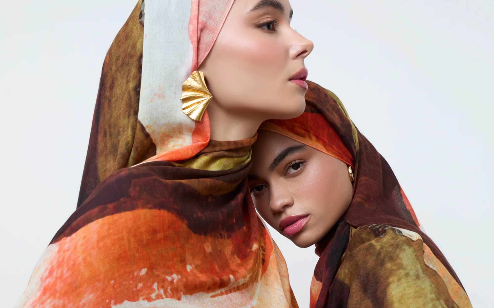 Two women wearing earthy-toned, painterly printed hijabs in rich rust, brown, and olive shades, positioned close together in a softly styled studio portrait.