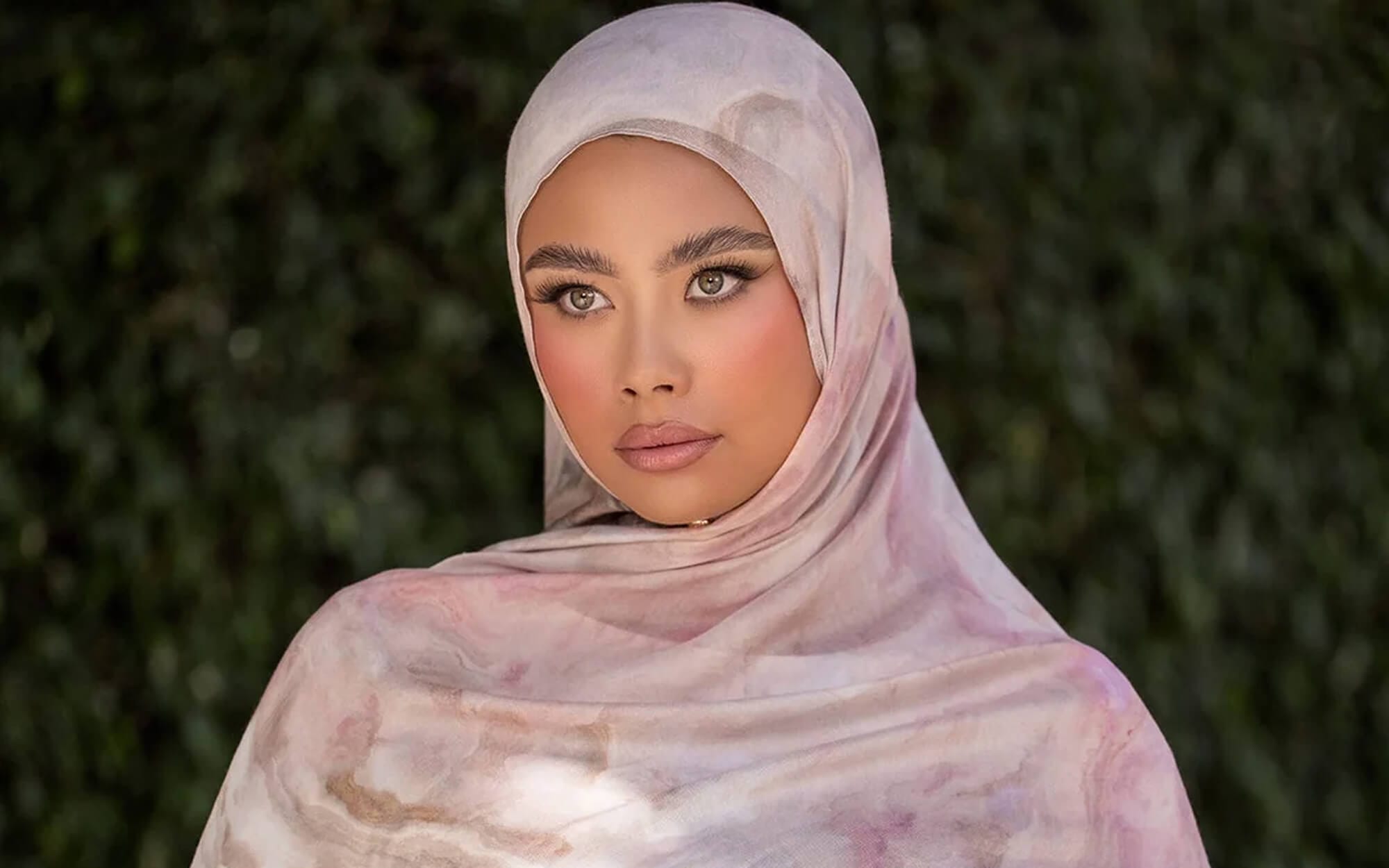A woman wearing a soft marble-print hijab in pink and beige tones, photographed against a green foliage background.