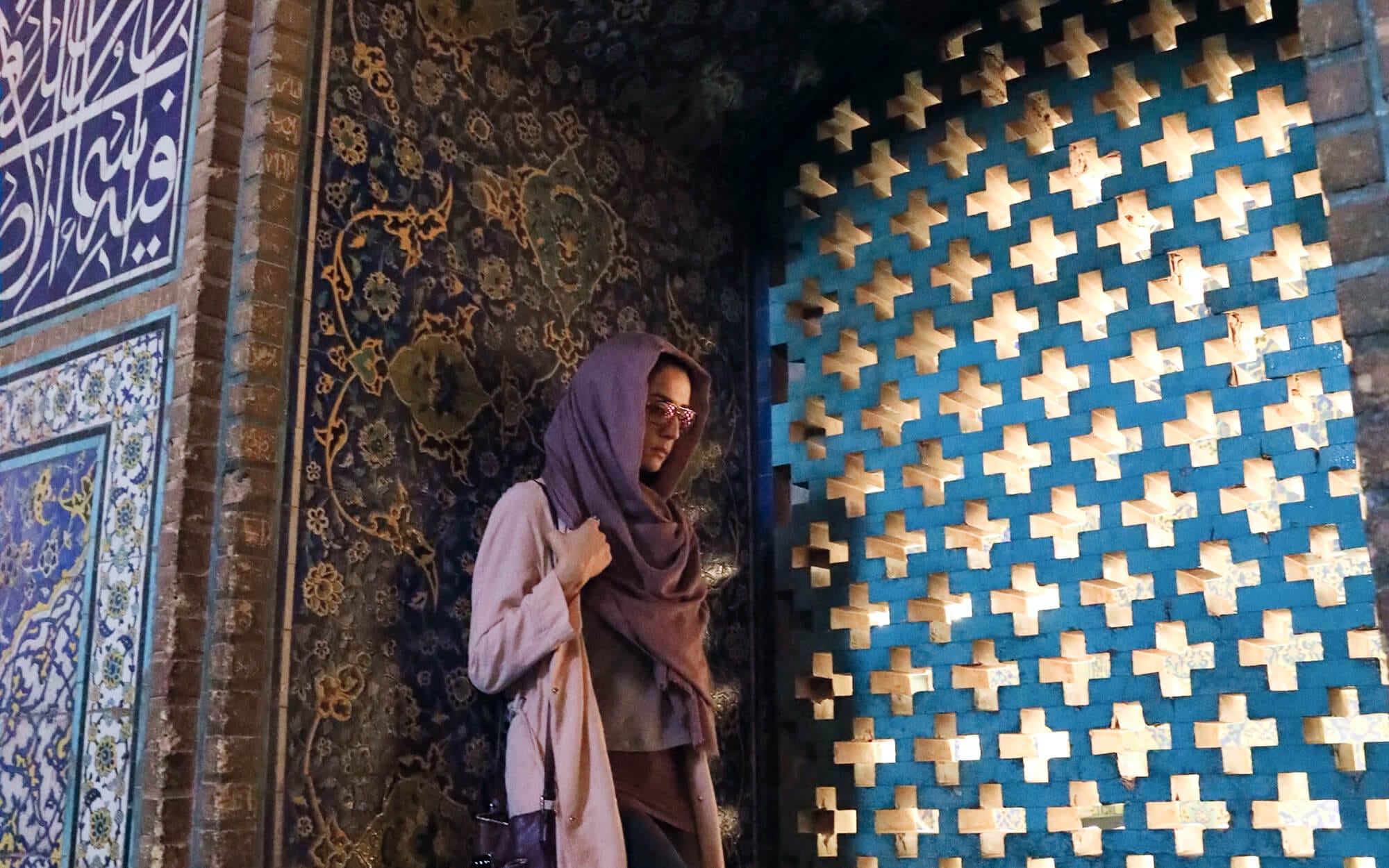 A person standing in a softly lit corner of Sheikh Lotfollah Mosque, beside a window filled with geometric star-shaped cutouts that filter warm light, with richly patterned blue-and-gold tiles covering the surrounding walls