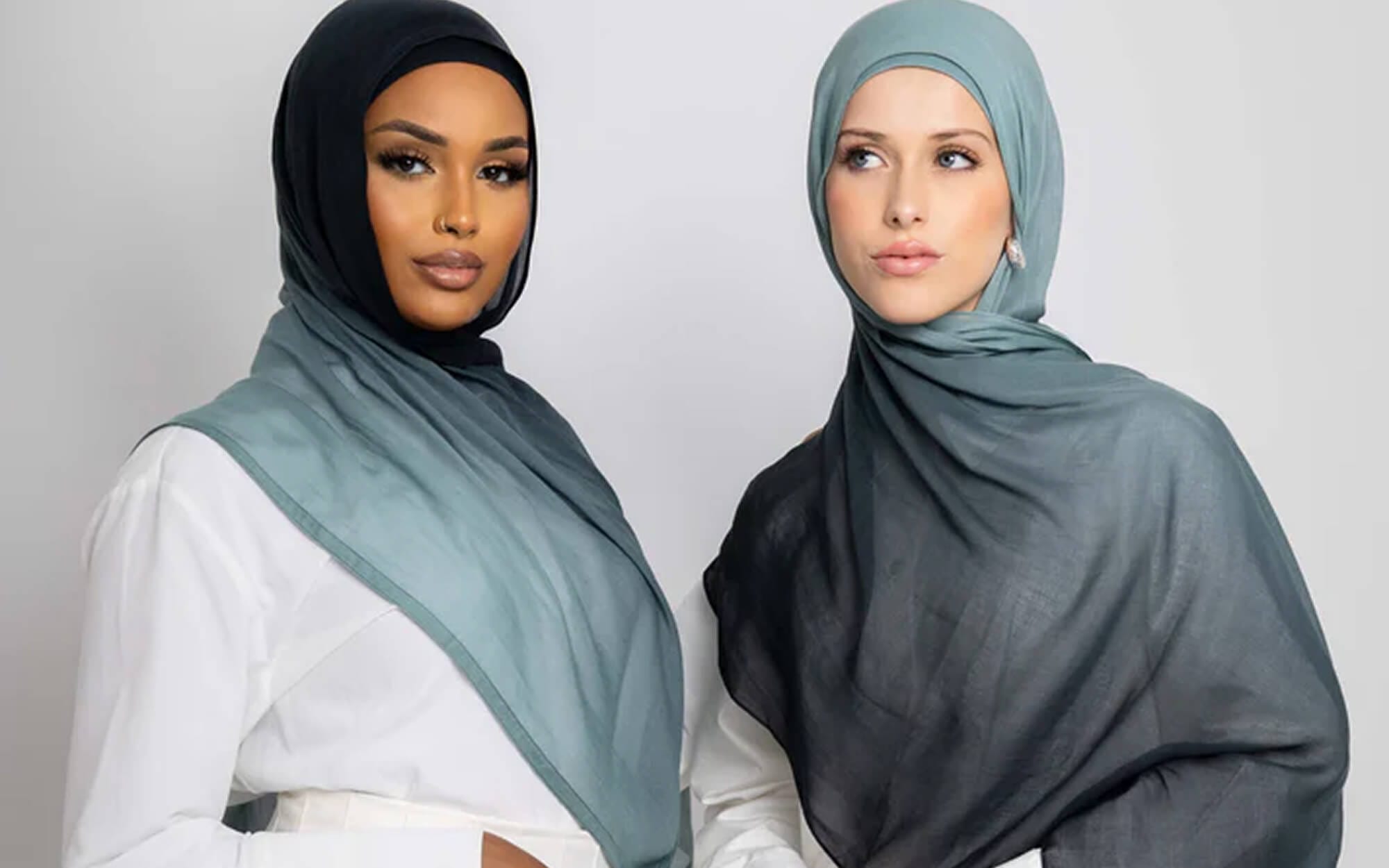 Two women wearing ombré hijabs in muted blue-green tones, one lighter and one darker, styled smoothly around their shoulders against a neutral background.