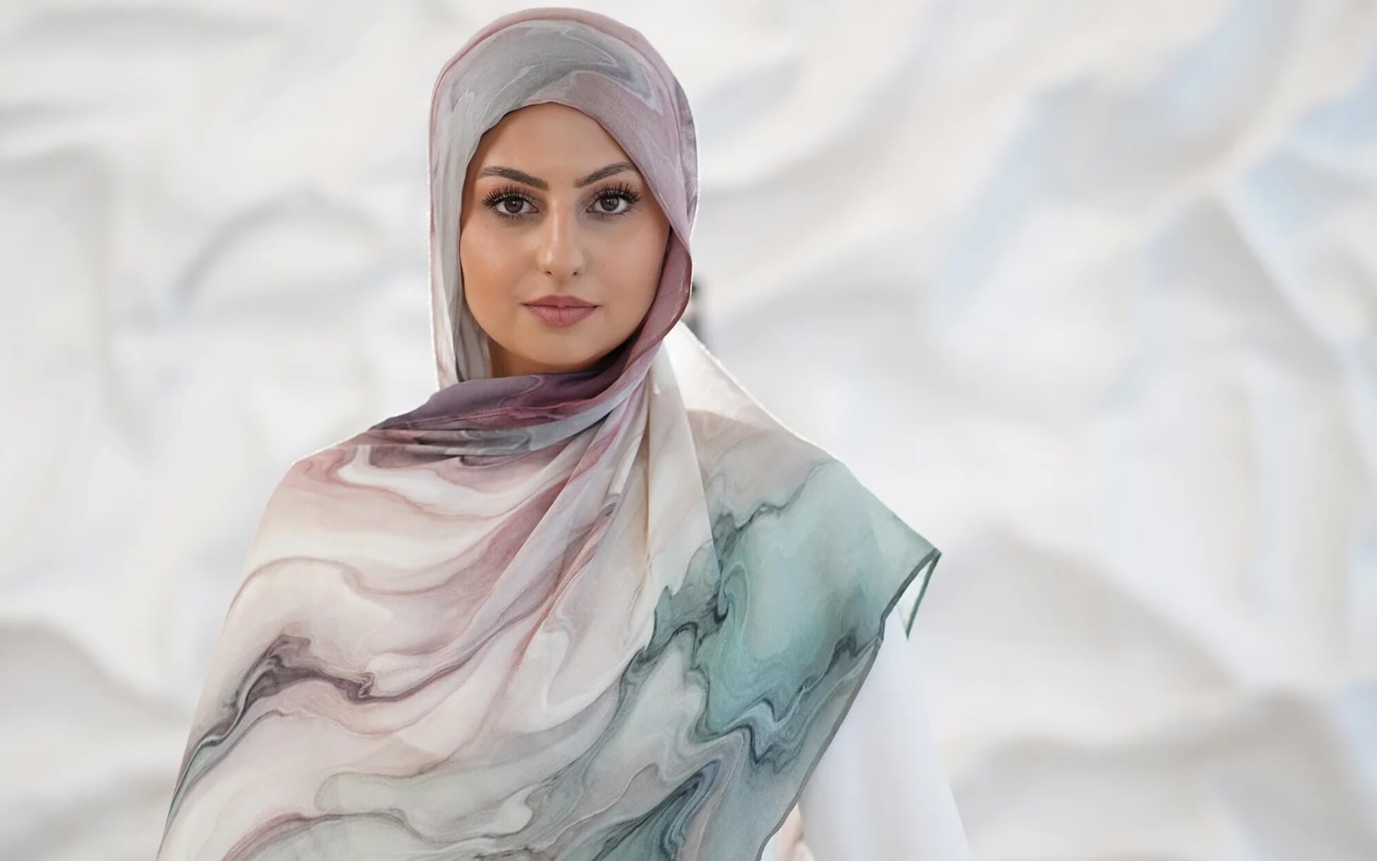 A woman wearing a flowing marble-print hijab in soft blush, mauve, and sage tones, standing in front of a textured light background.
