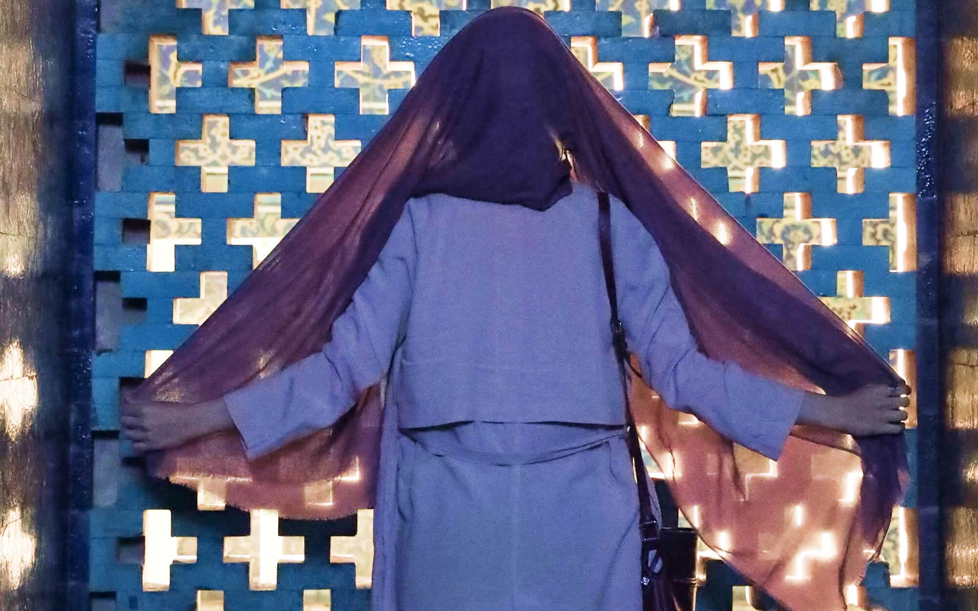 Person standing with back to the camera, holding a sheer purple shawl open in front of a blue-and-gold perforated geometric screen at Sheikh Lotfollah Mosque, with sunlight filtering through the pattern.