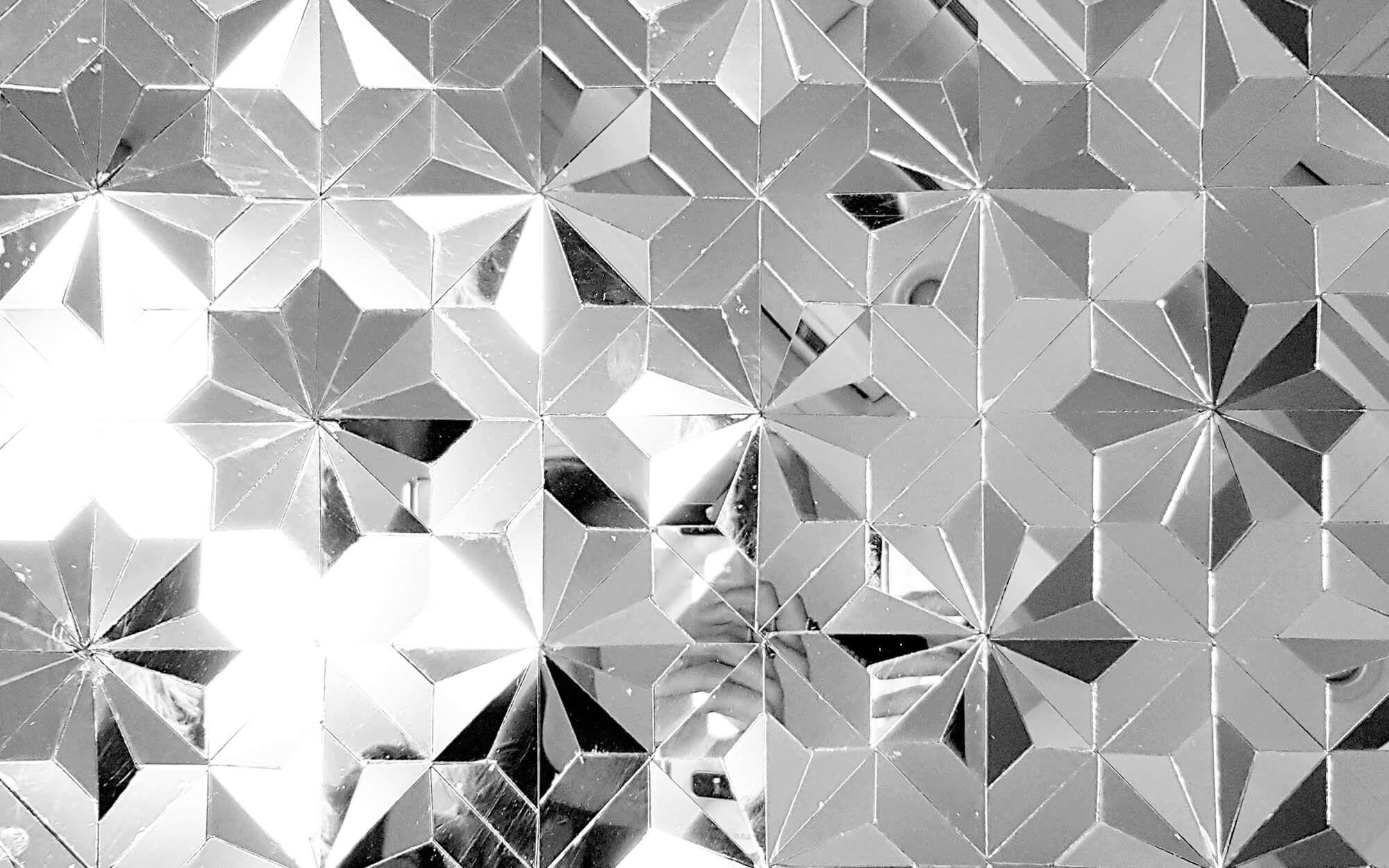Close-up of minimalist Āina-kāri (Persian mirror work) featuring a repeating geometric star pattern made from faceted mirror tiles that reflect and fragment light.