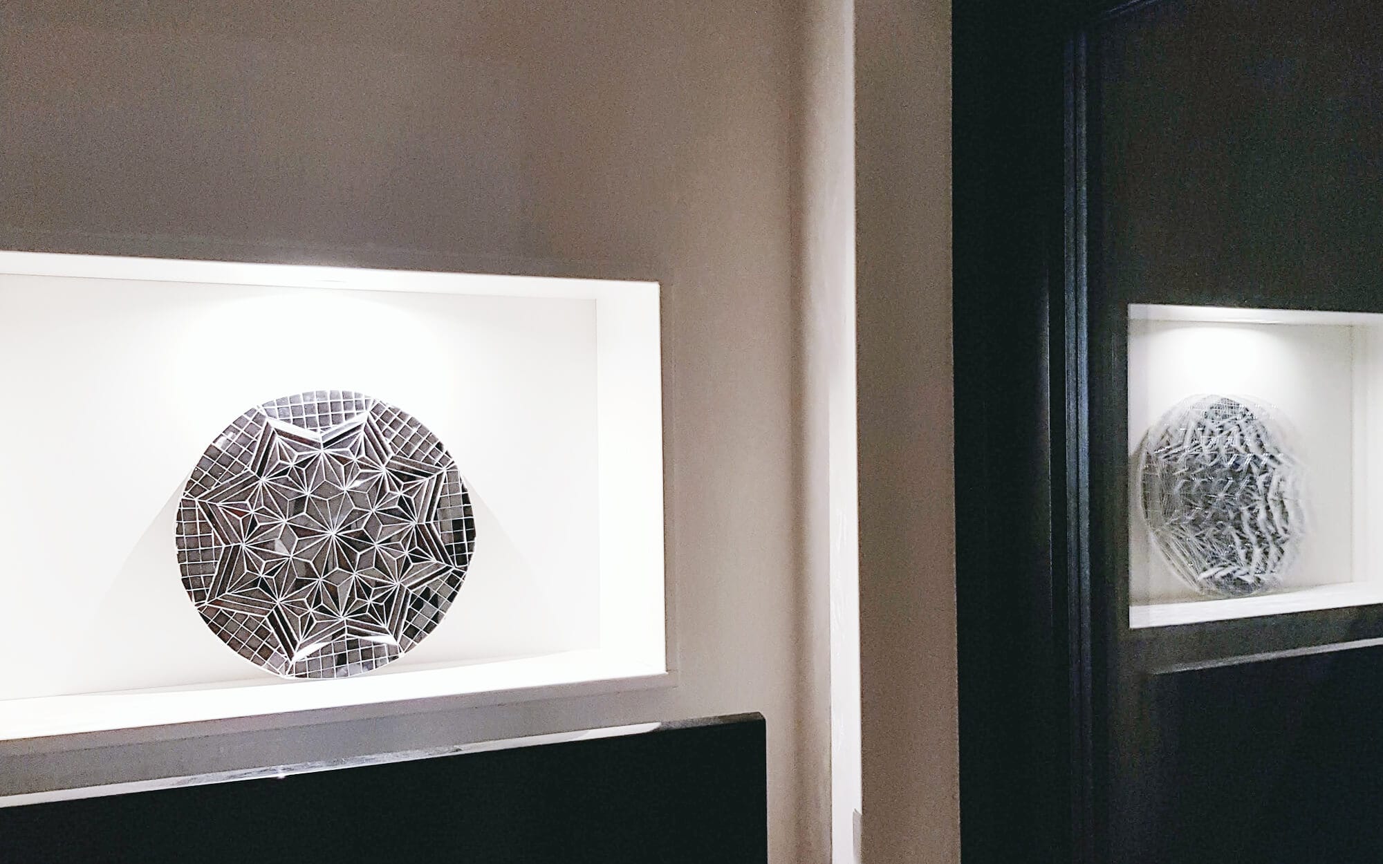 Minimal geometric mirror artwork displayed on a recessed wall shelf, softly lit within a white frame, with its reflection visible in a nearby mirror.