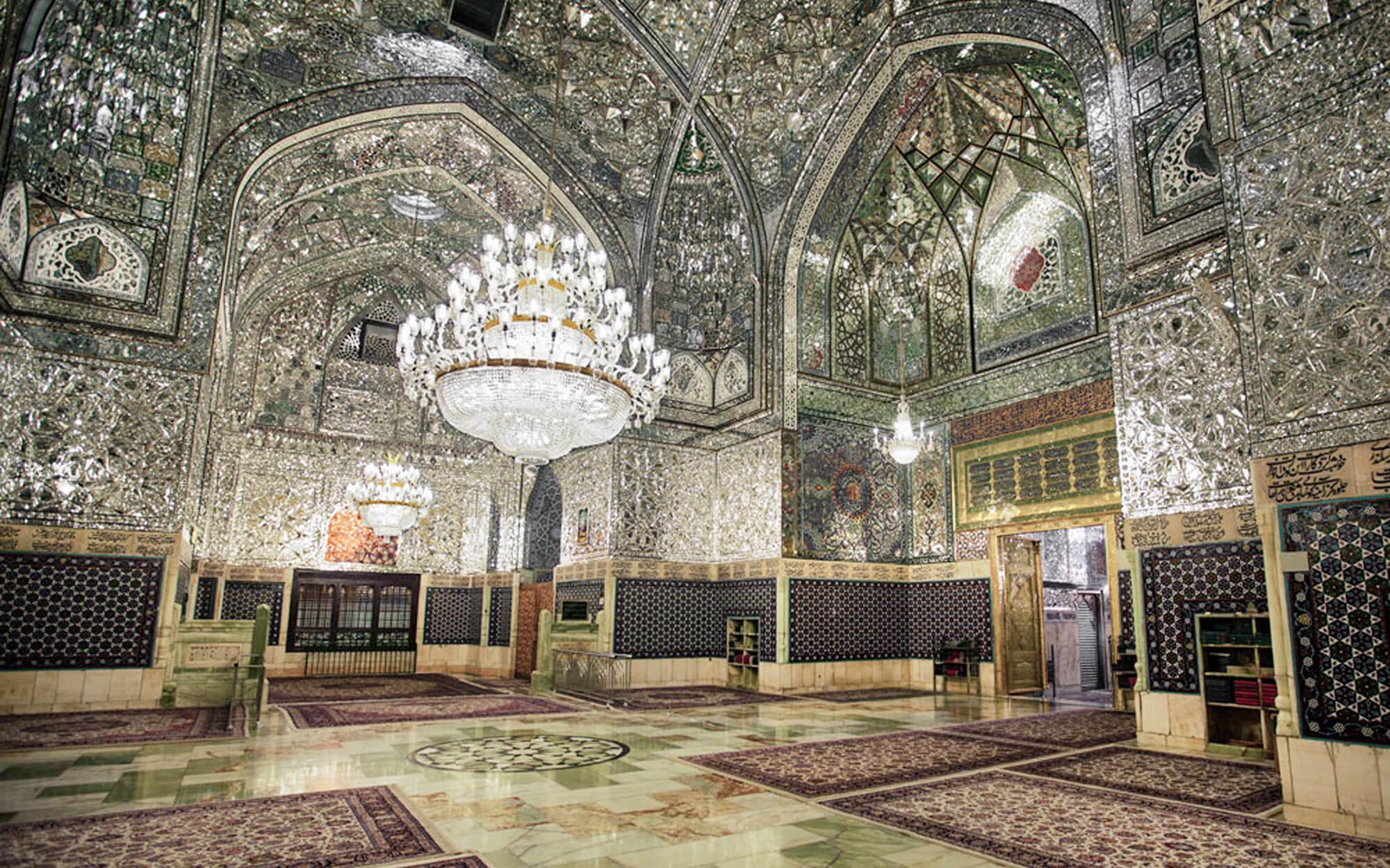 Interior of Imam Reza Shrine in Mashhad, showing walls and vaulted ceilings covered in intricate mirror mosaics, ornate stained-glass windows, and a large crystal chandelier above patterned marble floors and prayer carpets.