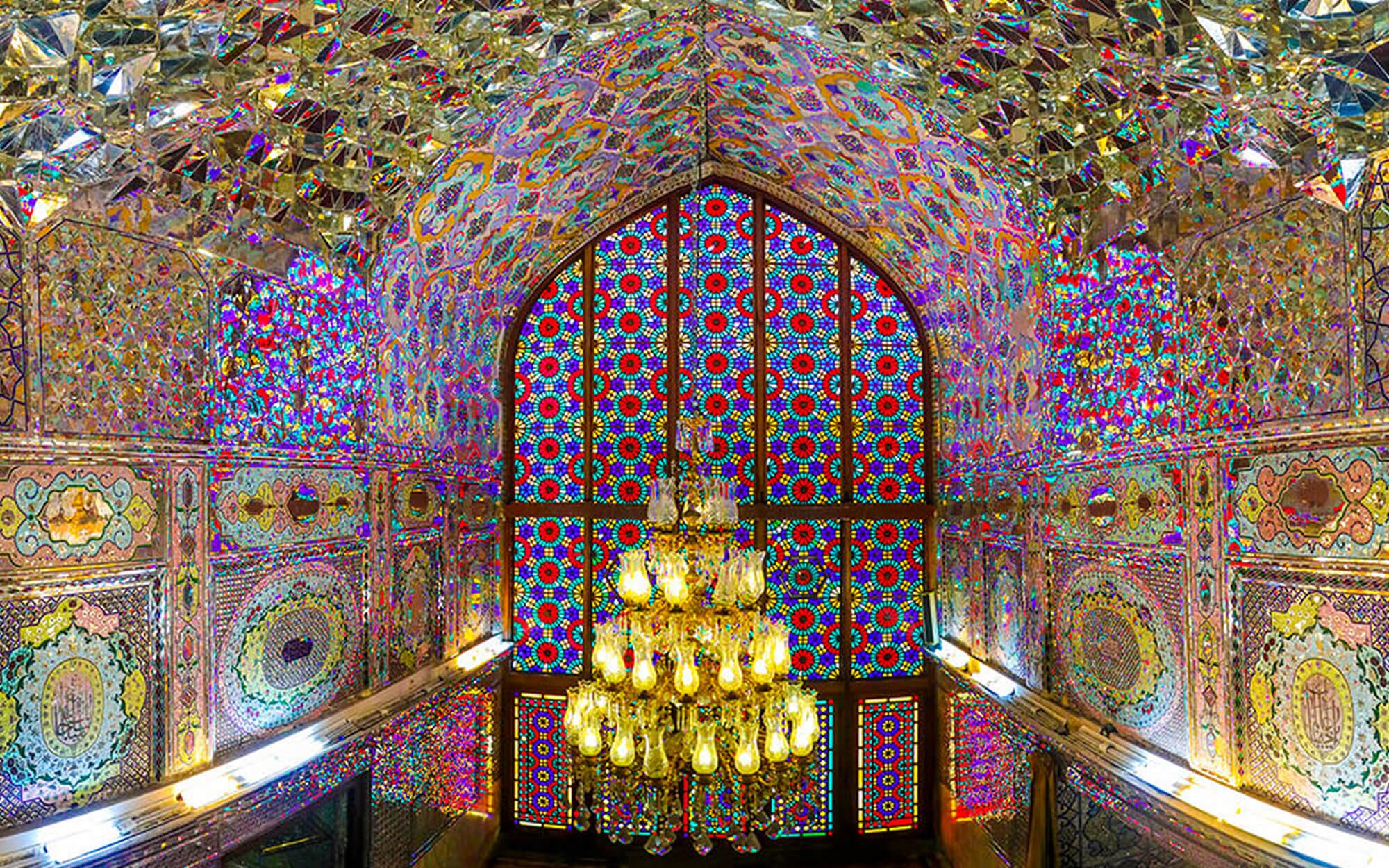Interior of Shah Cheragh in Shiraz, covered floor to ceiling in shimmering Āina-kāri mirror mosaics that reflect multicoloured light around a central stained-glass window and crystal chandelier.