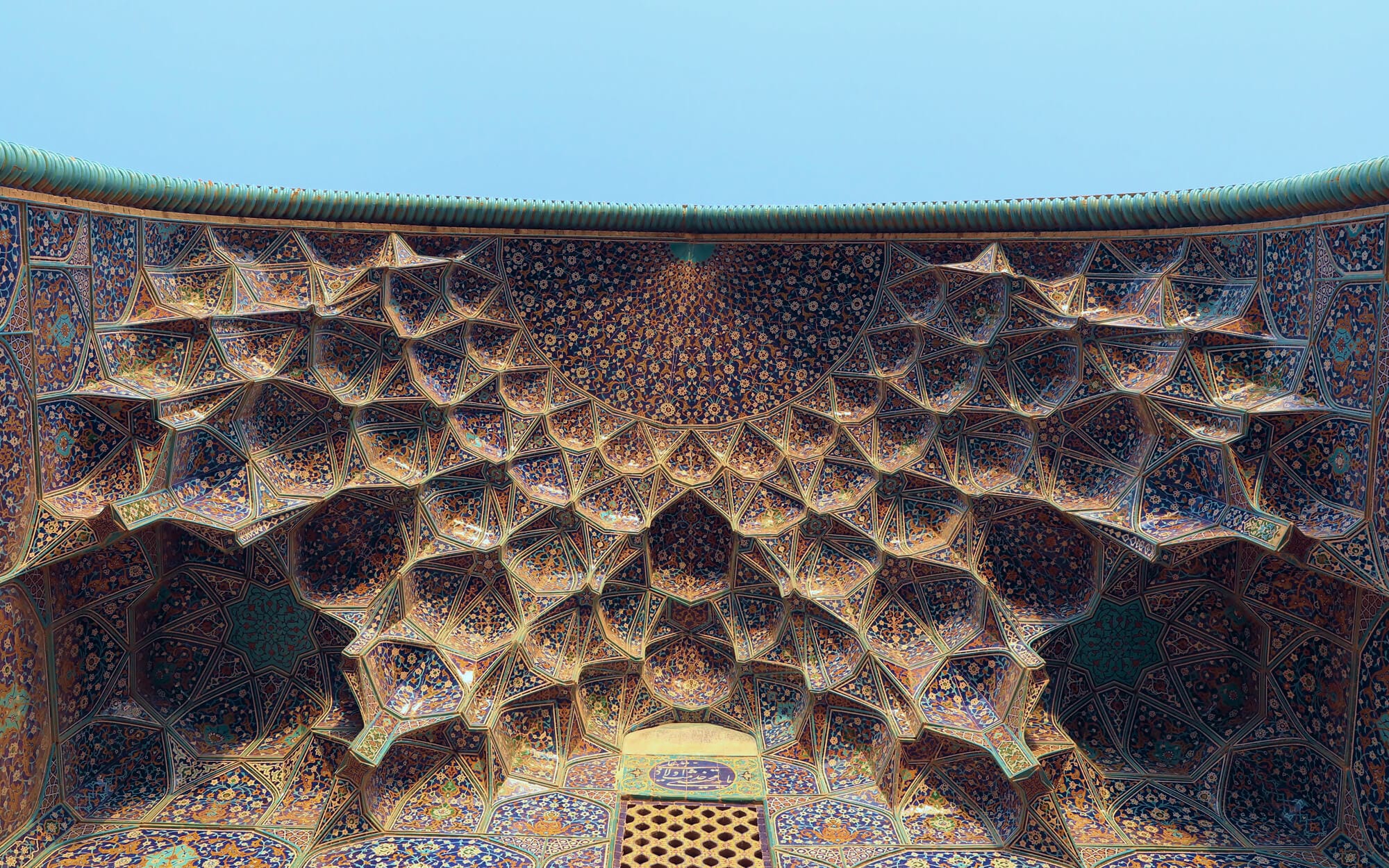Intricately layered muqarnas ceiling of Sheikh Lotfollah Mosque, formed by honeycomb-like geometric facets in deep blue, turquoise, and gold that appear to ripple across the dome under soft daylight.