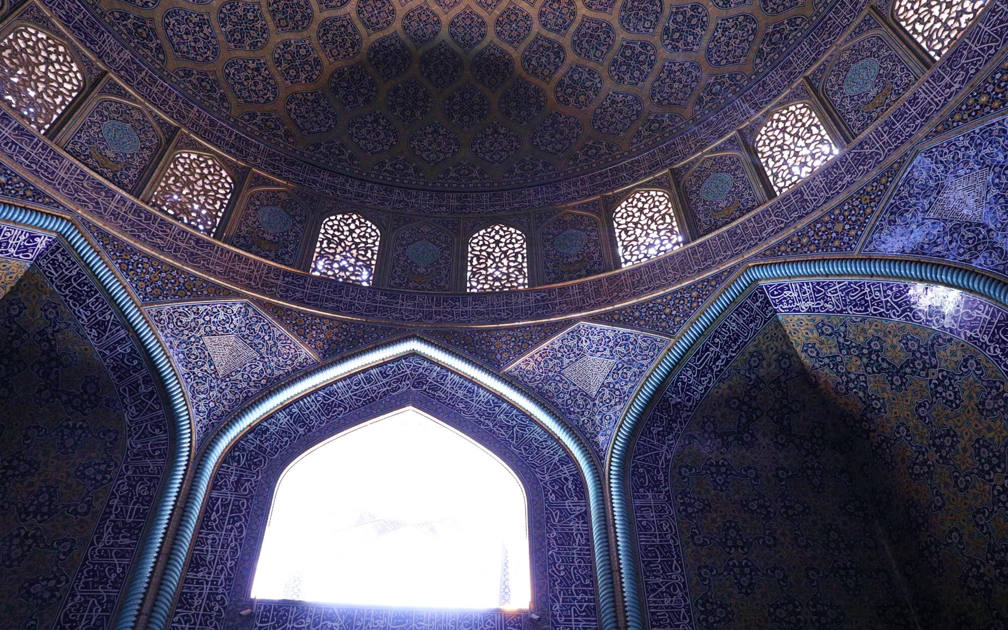 Interior dome of Sheikh Lotfollah Mosque, covered in intricate honeycomb-like geometric patterns, with carved lattice windows allowing shafts of light to spill across the richly tiled arches and walls.