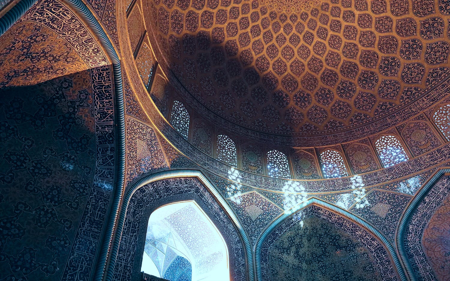 Interior dome of Sheikh Lotfollah Mosque, covered in intricate honeycomb-like geometric patterns, with carved lattice windows allowing shafts of light to spill across the richly tiled arches and walls.