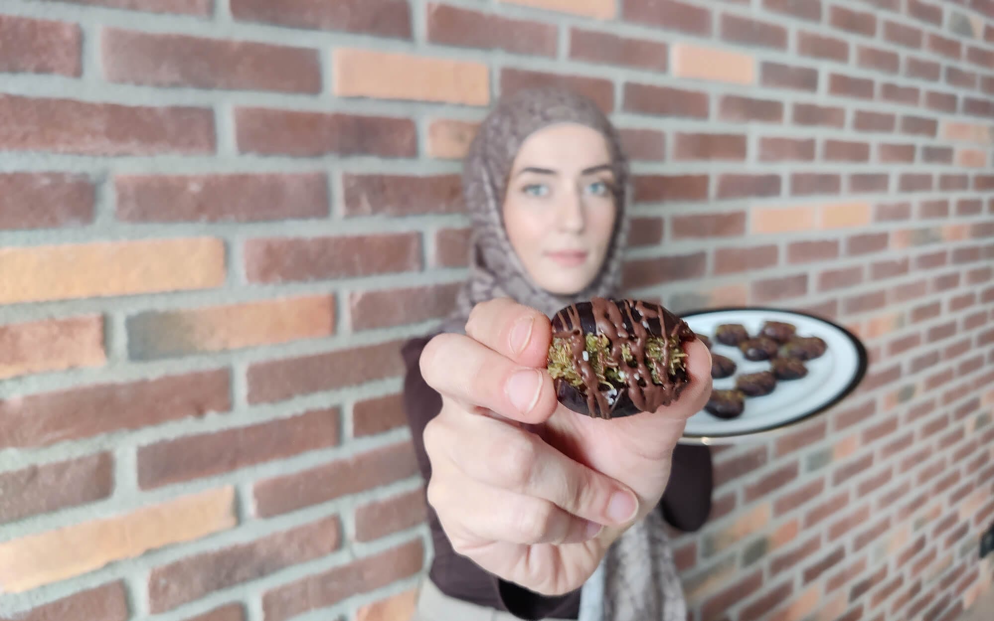 A hijabi woman holding up a homemade Dubai chocolate date toward the camera, with a plate of similar dates behind her against a brick wall.