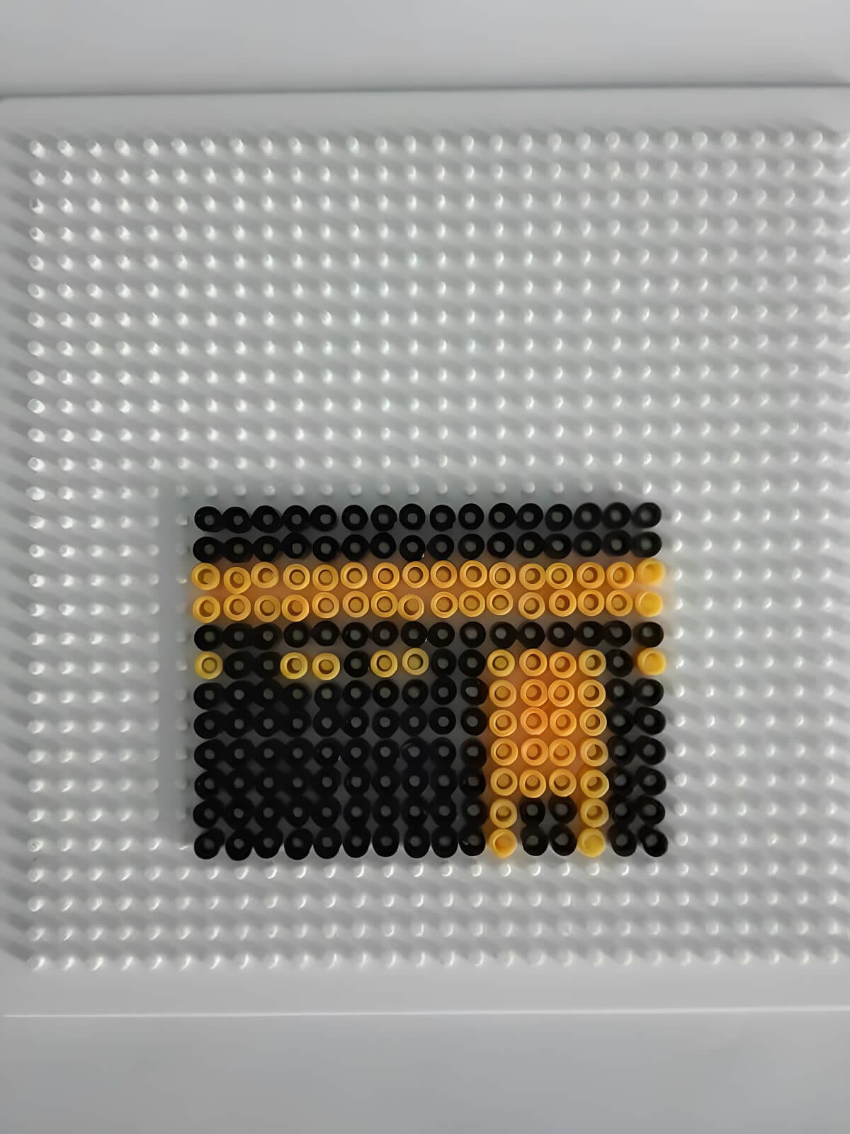 A pixel-style Kaaba made from black and gold Perler beads, with a simple gold “door” detail and a band across the top.