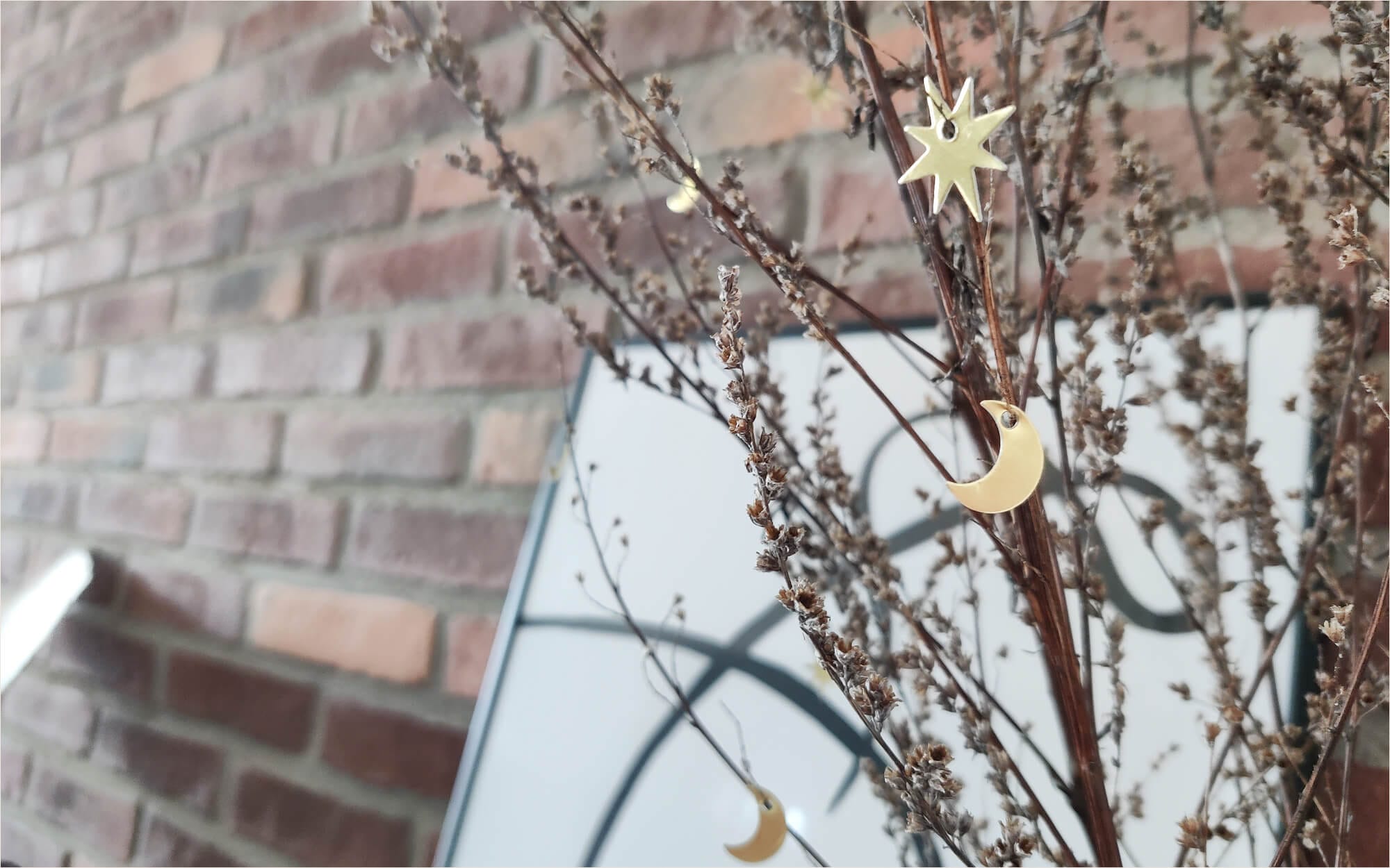 Ramadan Decor: Gold crescent moon and star ornaments hanging on dried branches in front of a framed minimalist line drawing against a brick wall.
