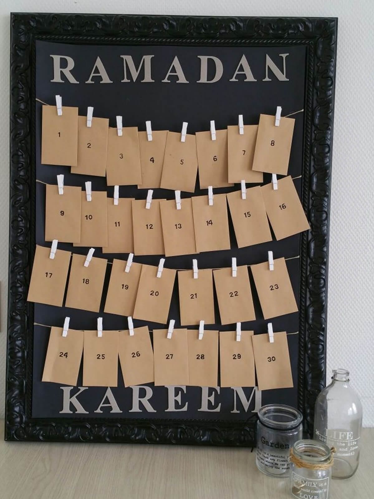 A framed “Ramadan Kareem” countdown display made from kraft paper envelopes clipped along three strings, numbered 1–30, hanging inside a large black decorative frame.