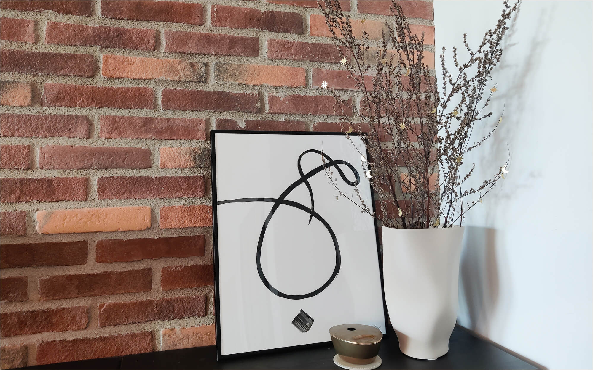 Minimal Ramadan branch decoration: dried branches adorned with small gold moon and star cutouts in a white vase, placed beside a framed minimalist calligraphy of the word “hubb” (love) in Arabic.