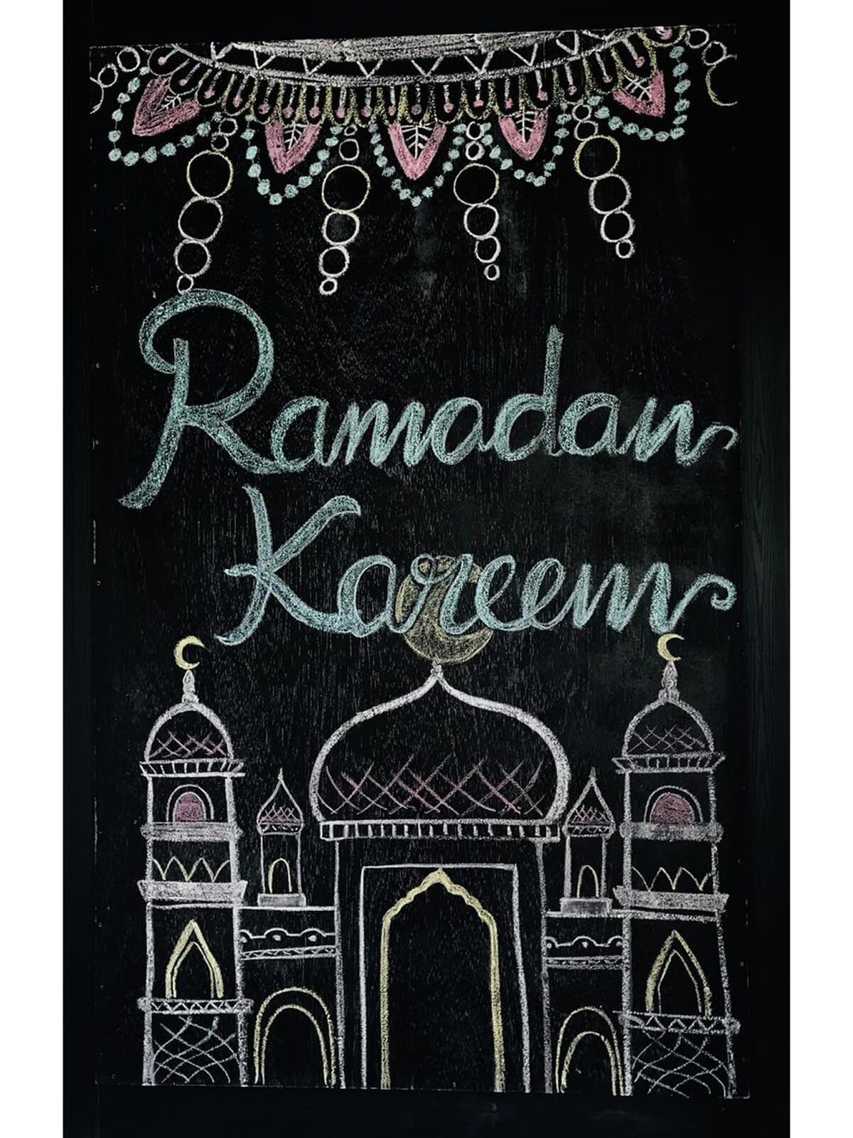 A hand-drawn “Ramadan Kareem” countdown board on a black chalkboard, decorated with pastel chalk lanterns, crescent details, and a mosque illustration at the bottom.