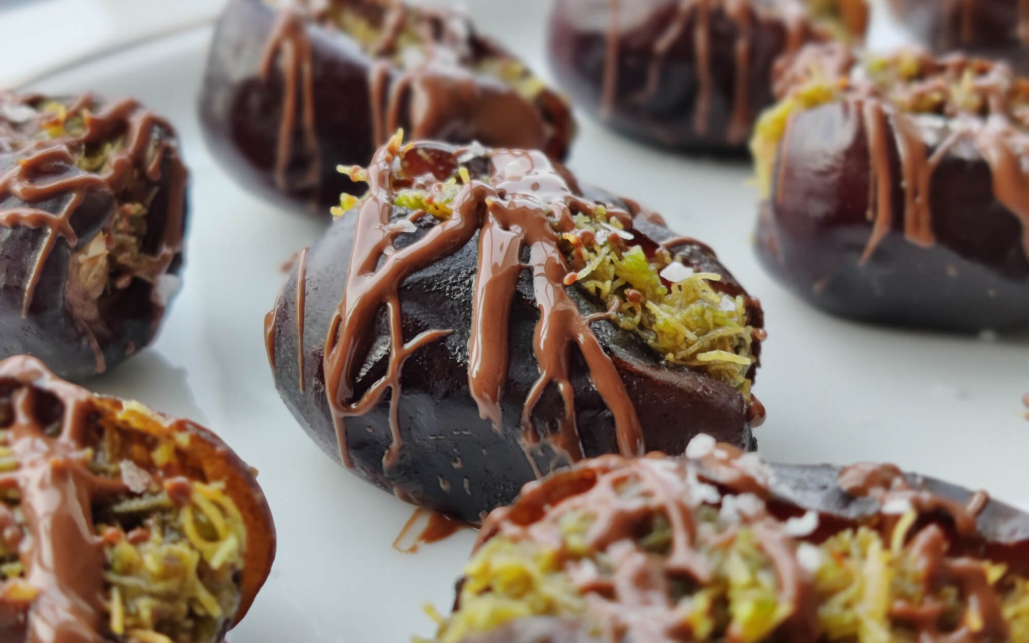 Dubai-style chocolate-stuffed dates filled with pistachio cream, Kataifi, and drizzled with melted chocolate on a white platter.