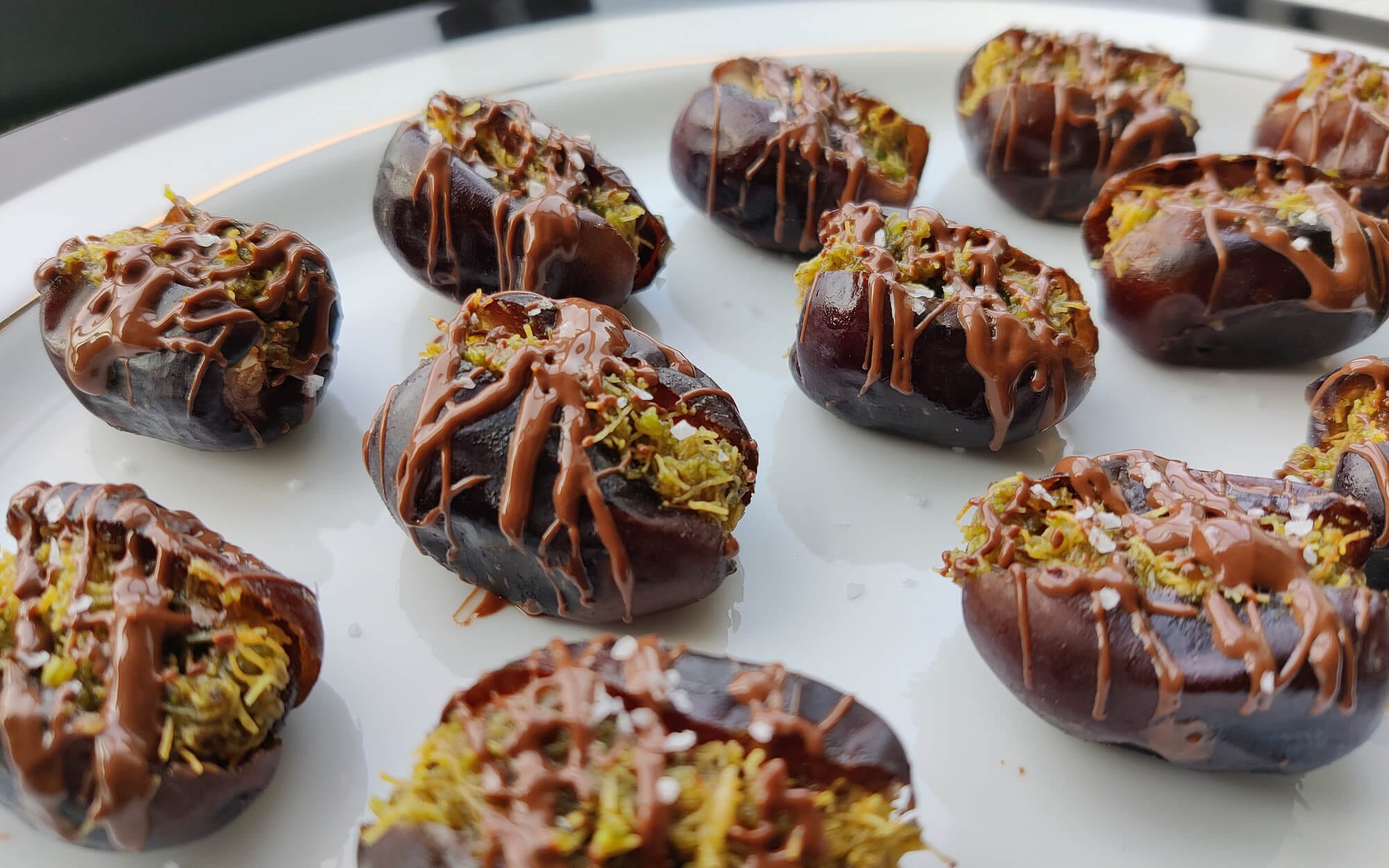Dubai-style chocolate-stuffed dates filled with pistachio cream, Kataifi, and drizzled with melted chocolate on a white platter.